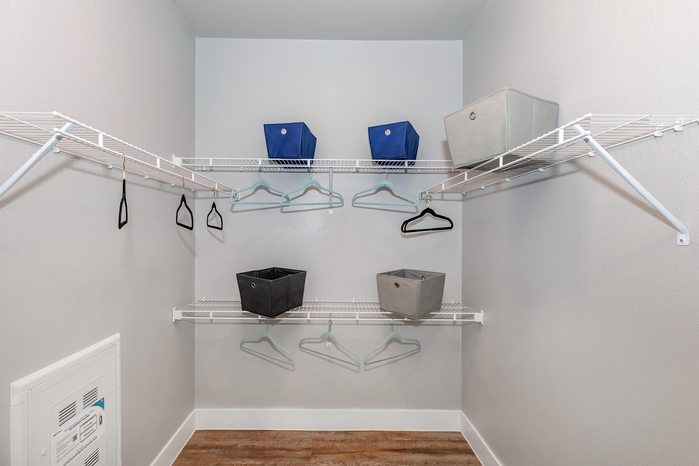 A well-organized closet featuring white wire shelving. Two blue fabric storage bins and one gray bin are placed on the shelves, while several black hangers hang from the lower shelf. The walls are painted light gray, and the floor is made of wood.
