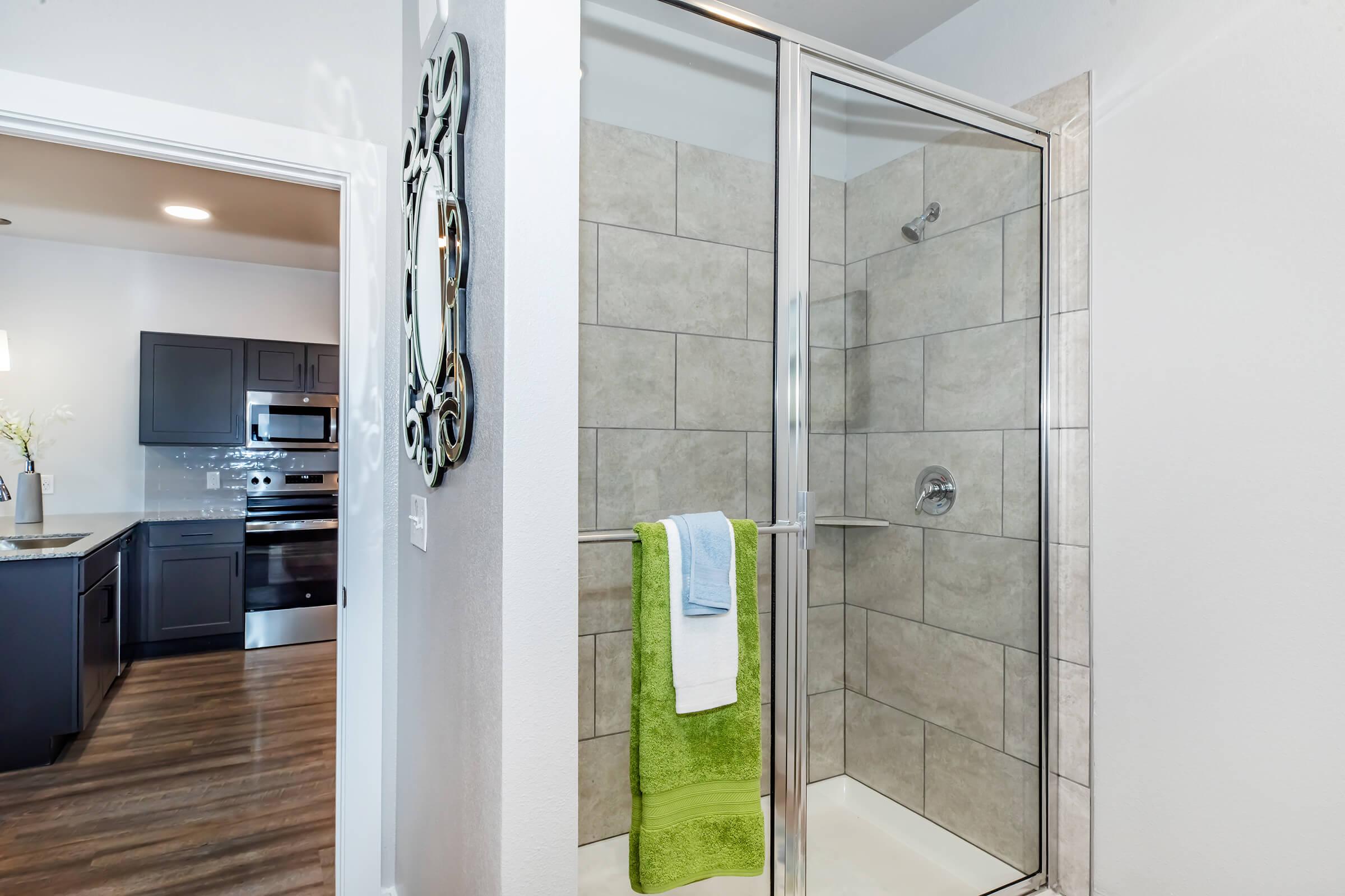 Modern bathroom featuring a glass shower enclosure with tile walls. A hanging green towel and two smaller towels in blue and white are visible. The background shows a glimpse of a kitchen with stainless steel appliances and dark cabinetry. The flooring is wood-like laminate.