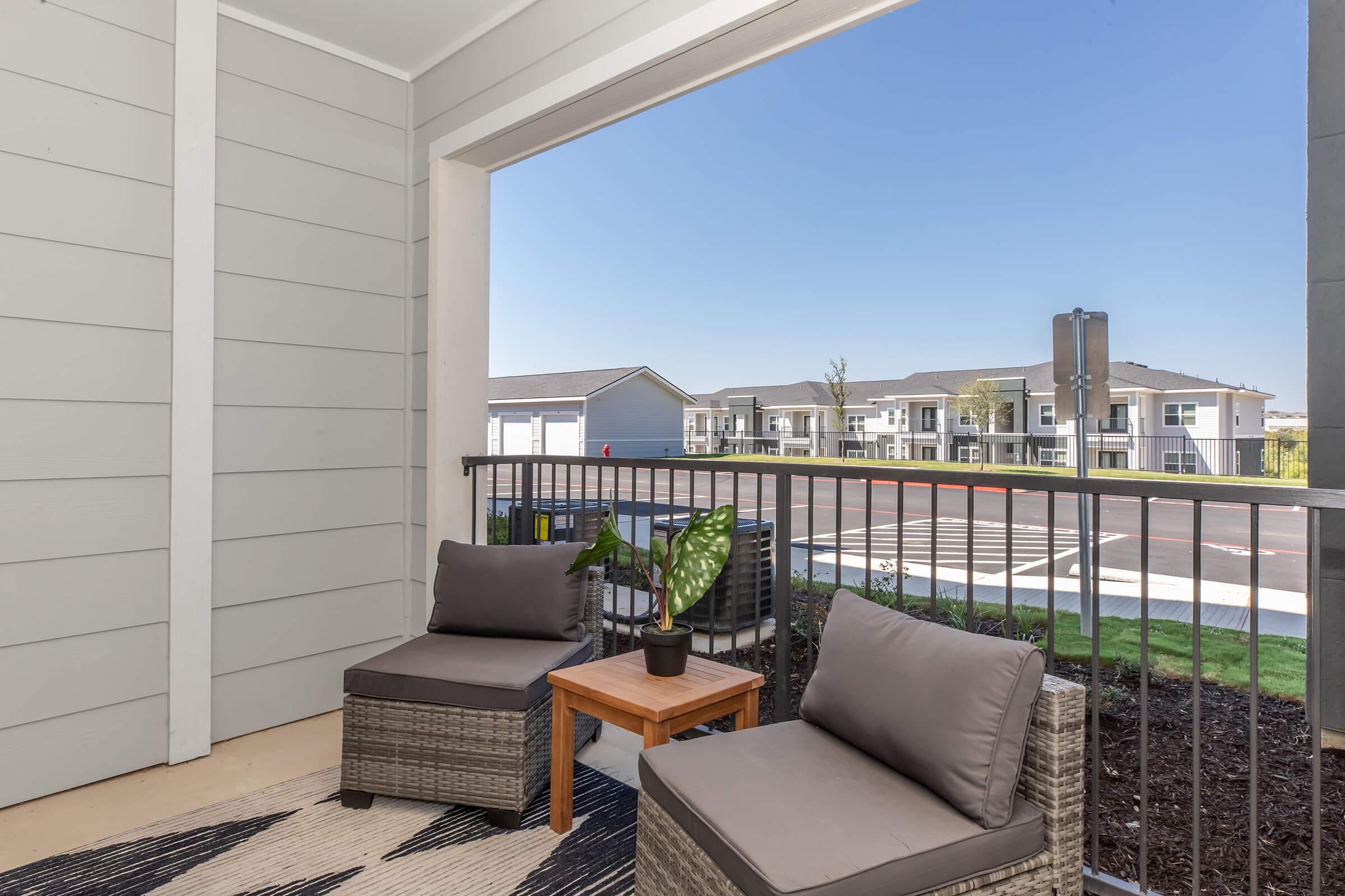 A cozy balcony with two lounge chairs and a small wooden table featuring a potted plant. The view includes a well-maintained apartment complex with green landscaping and clear blue skies. The setting is inviting and suitable for relaxation.