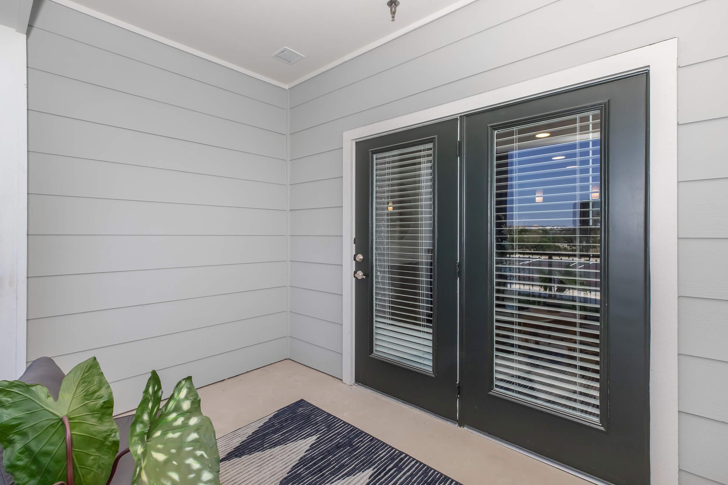 A modern entryway featuring double glass doors with horizontal blinds, framed by gray siding. The floor is beige, and a decorative area rug is visible. A green leafy houseplant adds a touch of nature, enhancing the inviting atmosphere of the space.
