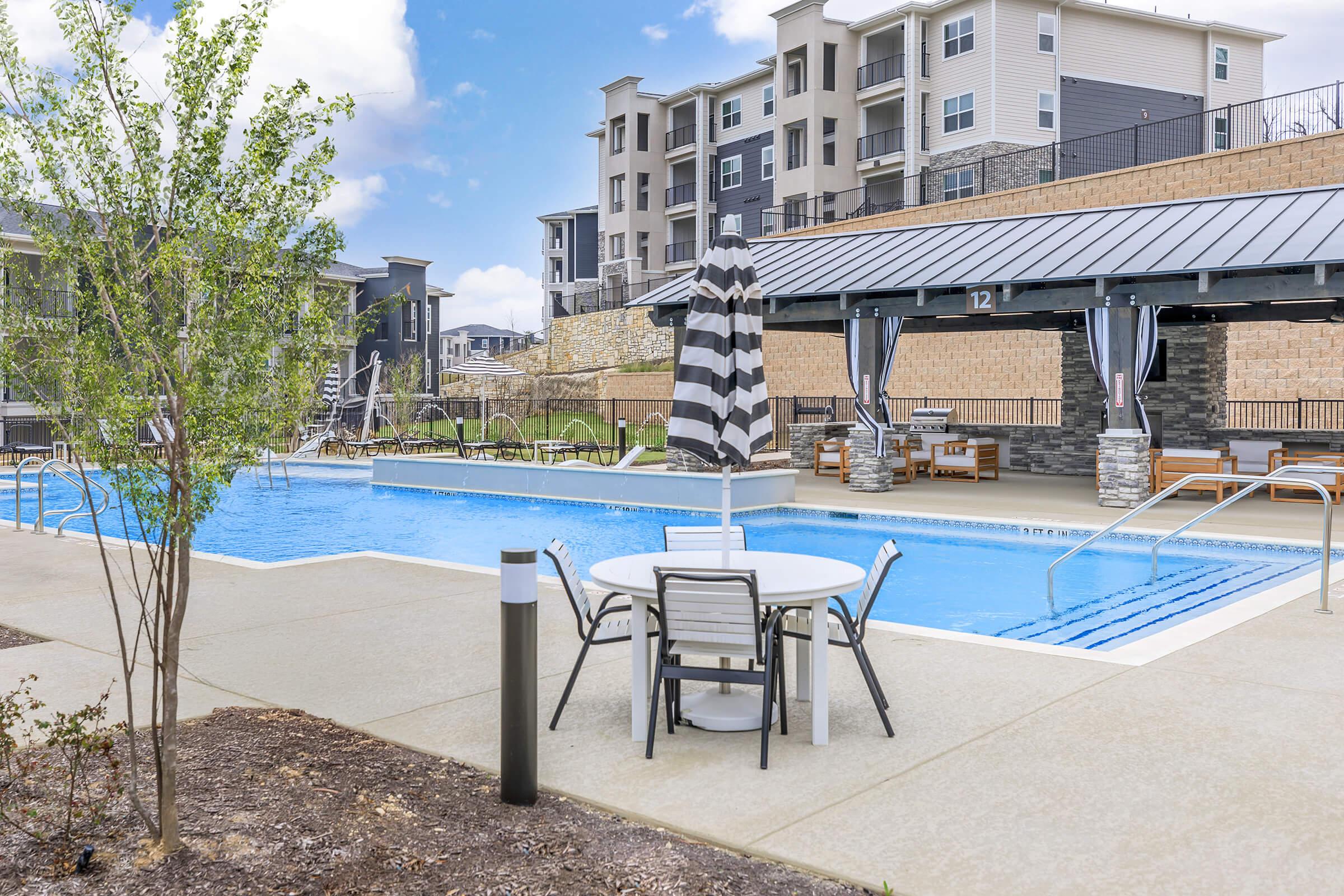 A clear blue swimming pool surrounded by lounge chairs and tables with umbrellas, set in a residential area. Modern apartment buildings are visible in the background, and landscaping features green bushes and a concrete pathway. The sky is partly cloudy, adding to the inviting atmosphere.