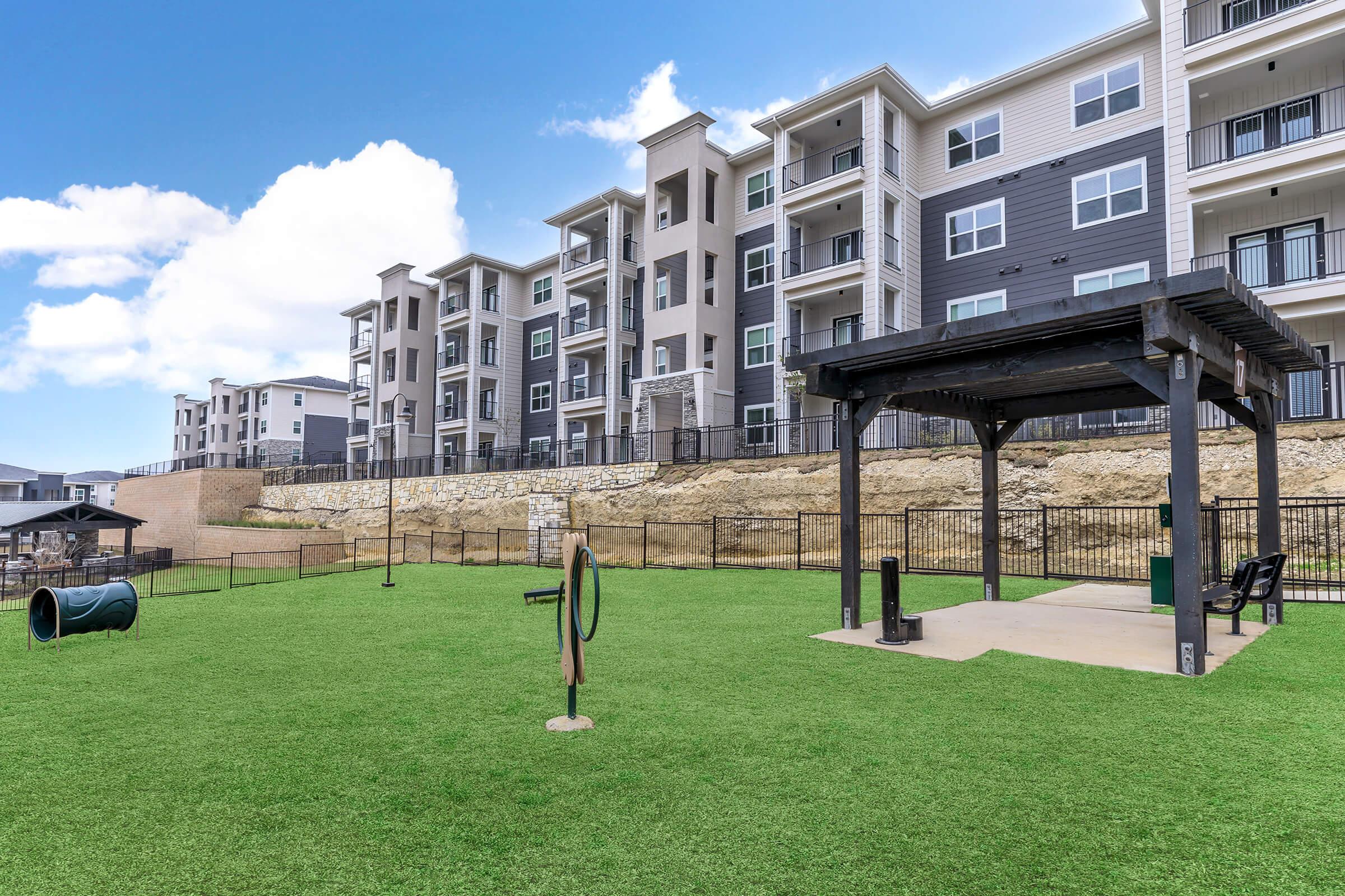 A modern residential apartment complex featuring multiple three-story buildings, with balconies and large windows. In the foreground, a spacious, green lawn includes a dog park with agility equipment, a covered seating area, and a play tunnel for pets, surrounded by a black metal fence.