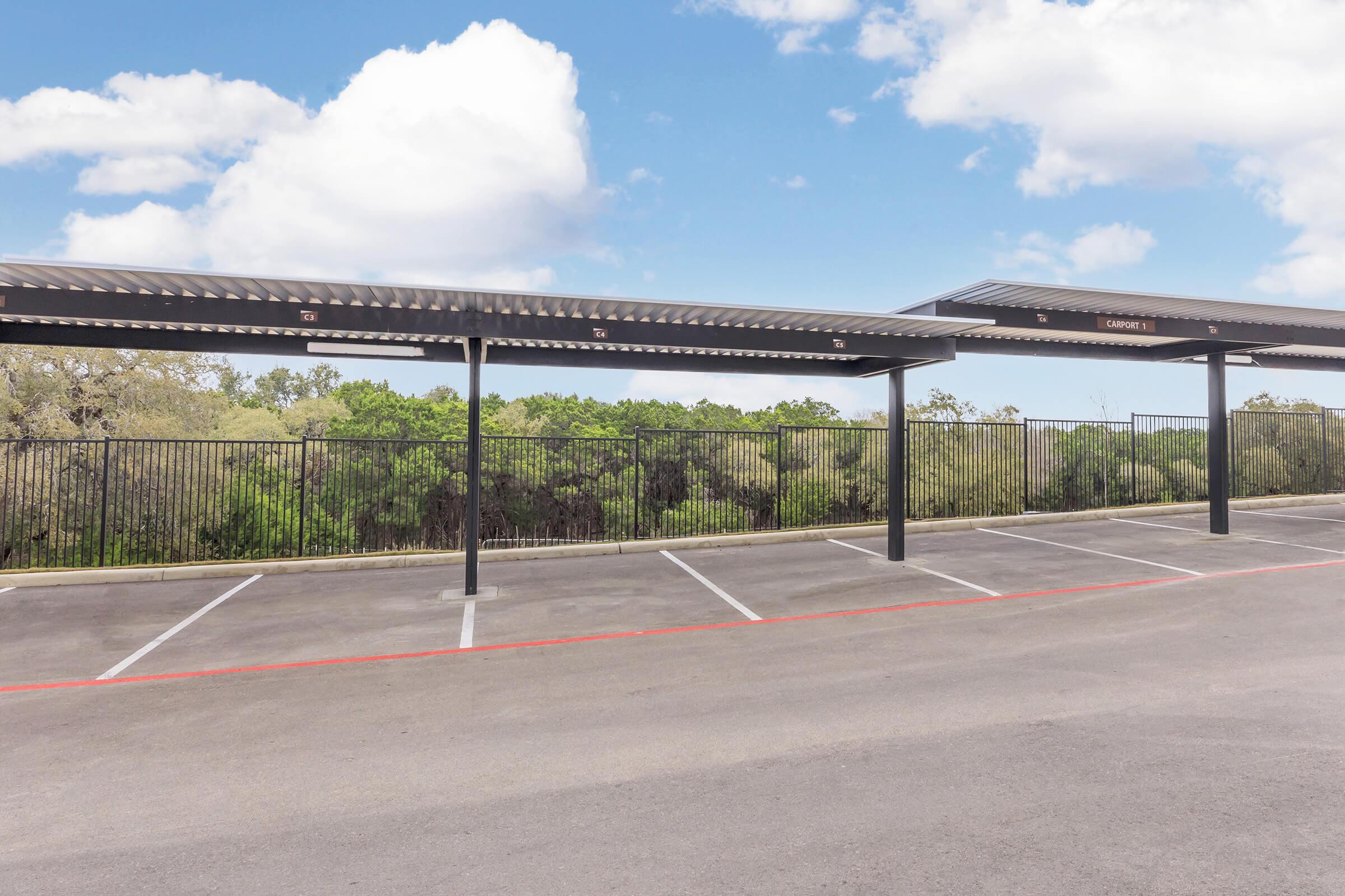 A parking lot with several empty spaces and shaded canopies. In the background, green trees and a blue sky with scattered clouds are visible.