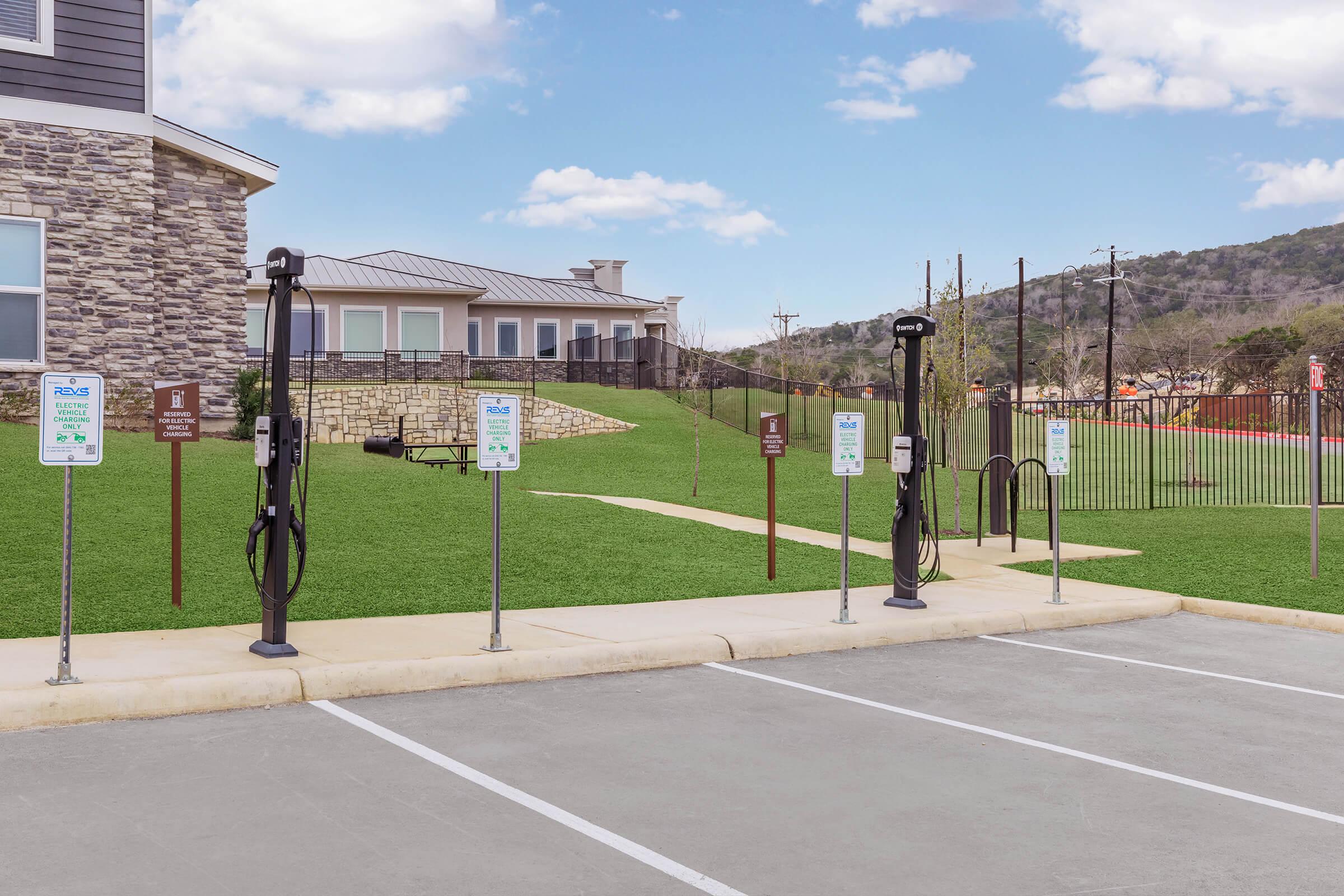 Electric vehicle charging stations are set up in a well-maintained parking area with green grass and a fence in the background. Signs indicate charging station usage and rules. The setting is sunny with a few clouds in the sky, and a building is visible to the left.