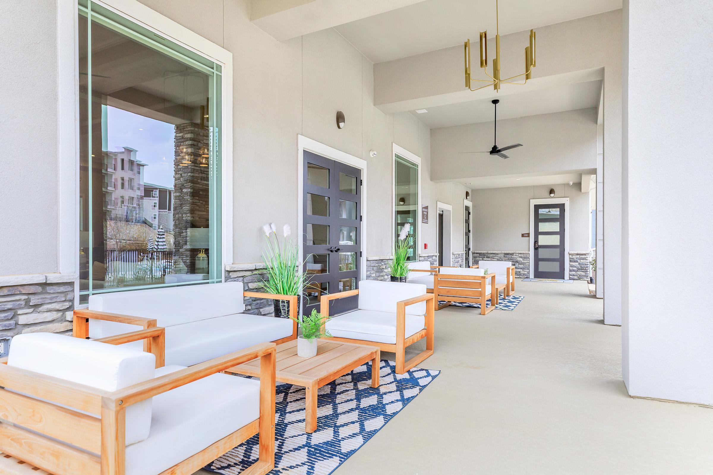 A modern outdoor seating area featuring white couches and wooden coffee tables on a light-colored floor. The space is decorated with potted plants and a patterned blue rug, surrounded by large windows. A stylish light fixture hangs from the ceiling, enhancing the contemporary aesthetic.