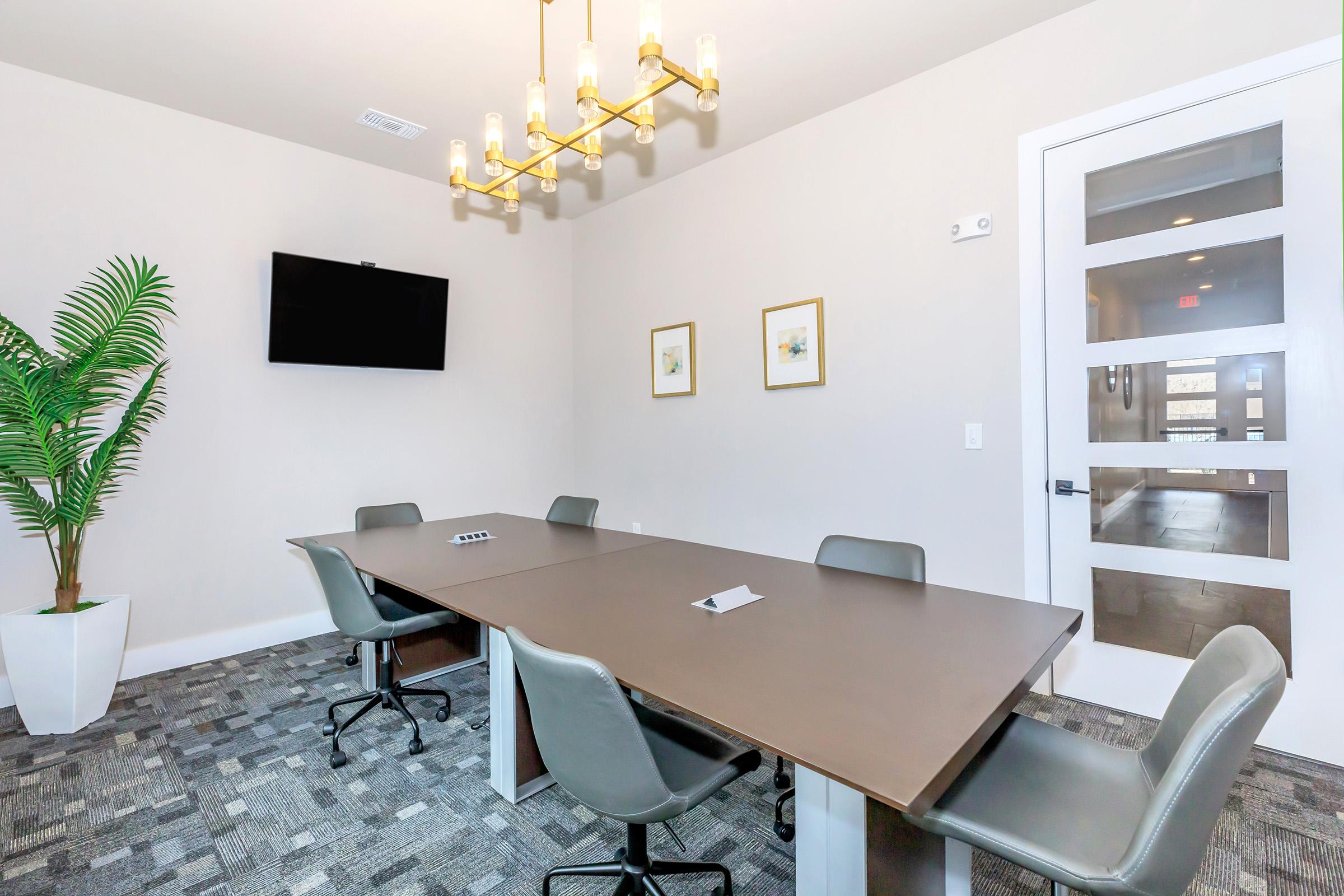A modern conference room featuring a long table surrounded by gray chairs, a potted plant, a TV mounted on the wall, and two framed pictures. A stylish chandelier hangs from the ceiling, and large windows provide natural light. The decor is sleek and professional, suitable for meetings.