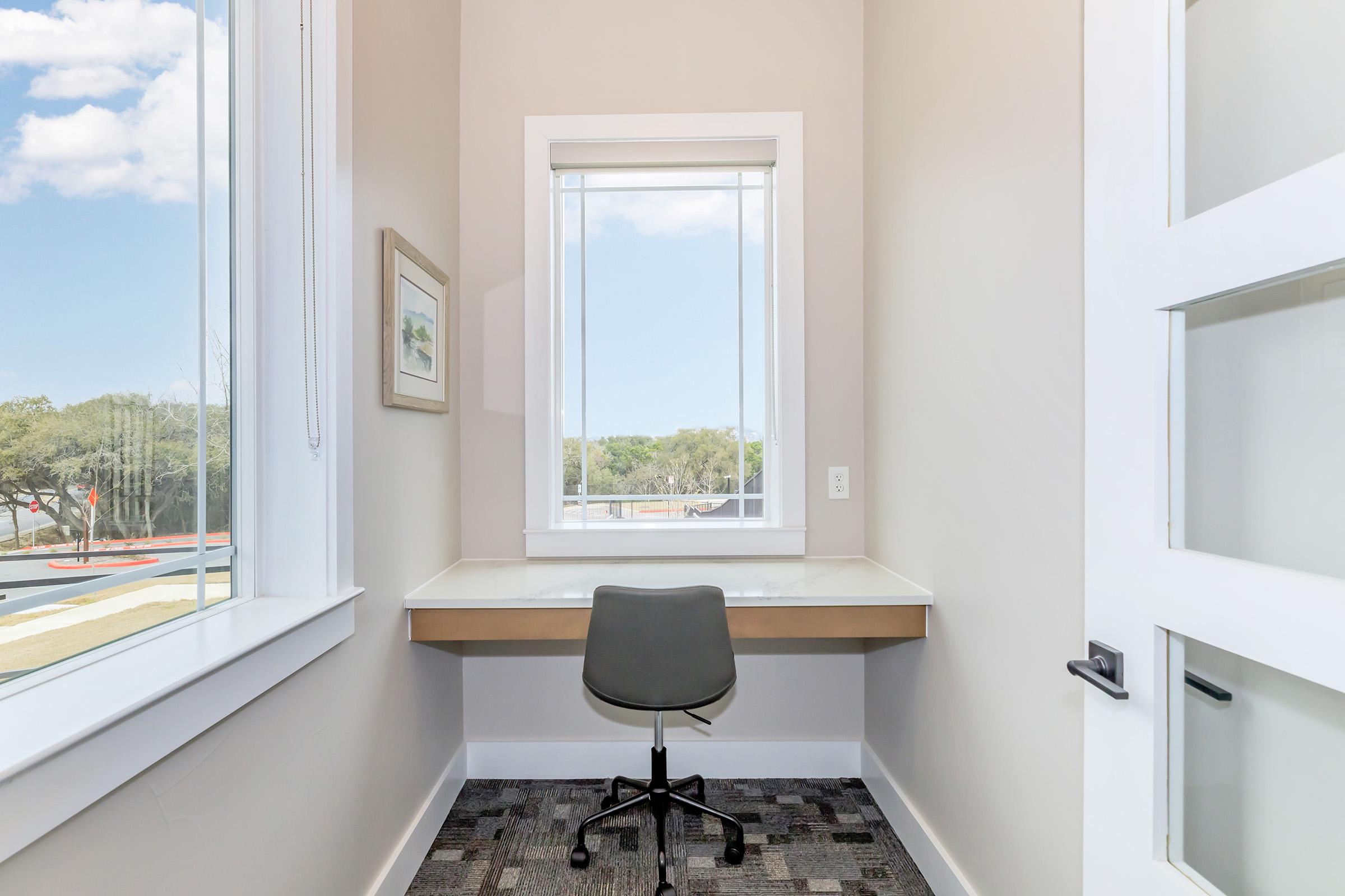 A small, bright workspace with a minimalistic design featuring a desk and a black office chair. Large windows provide natural light and a view of greenery outside. The walls are painted in neutral tones, and there's a framed picture on the wall, adding a touch of decor.