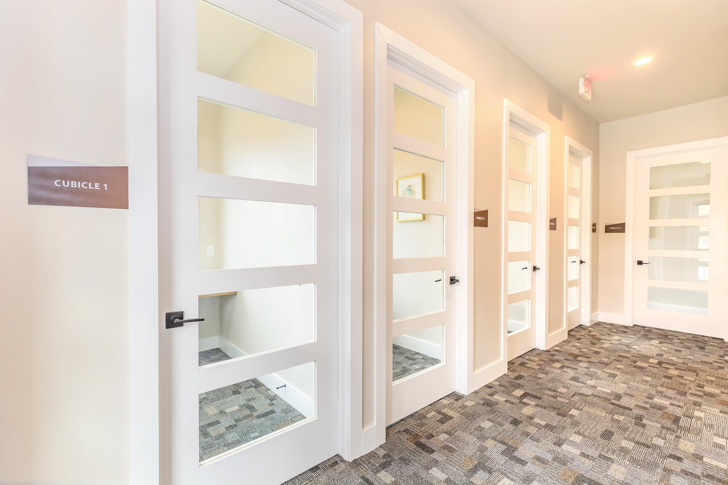A hallway featuring several white cubicle doors with glass panels. Each door is labeled with a number, and there is a modern carpet covering the floor. The walls are painted in a light color, creating a bright and clean atmosphere. Soft lighting enhances the professional look of the space.