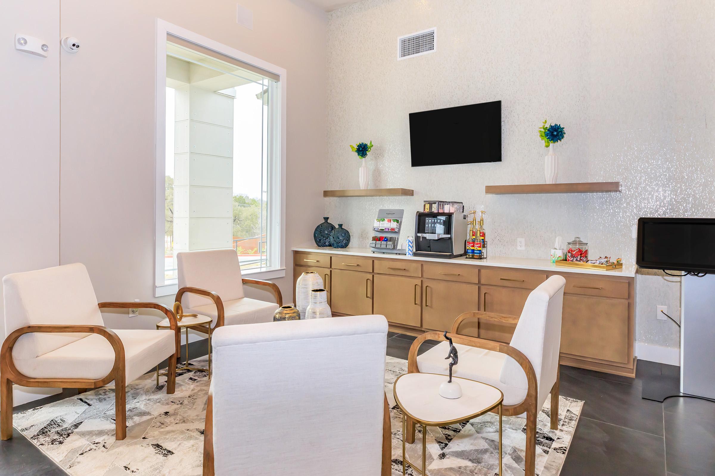 Cozy lounge area featuring four white chairs arranged around a small table, a television mounted on the wall, and a stylish sideboard displaying snacks and beverages. Large windows allow natural light, complemented by decorative vases with flowers on the shelves. The flooring is dark and modern.