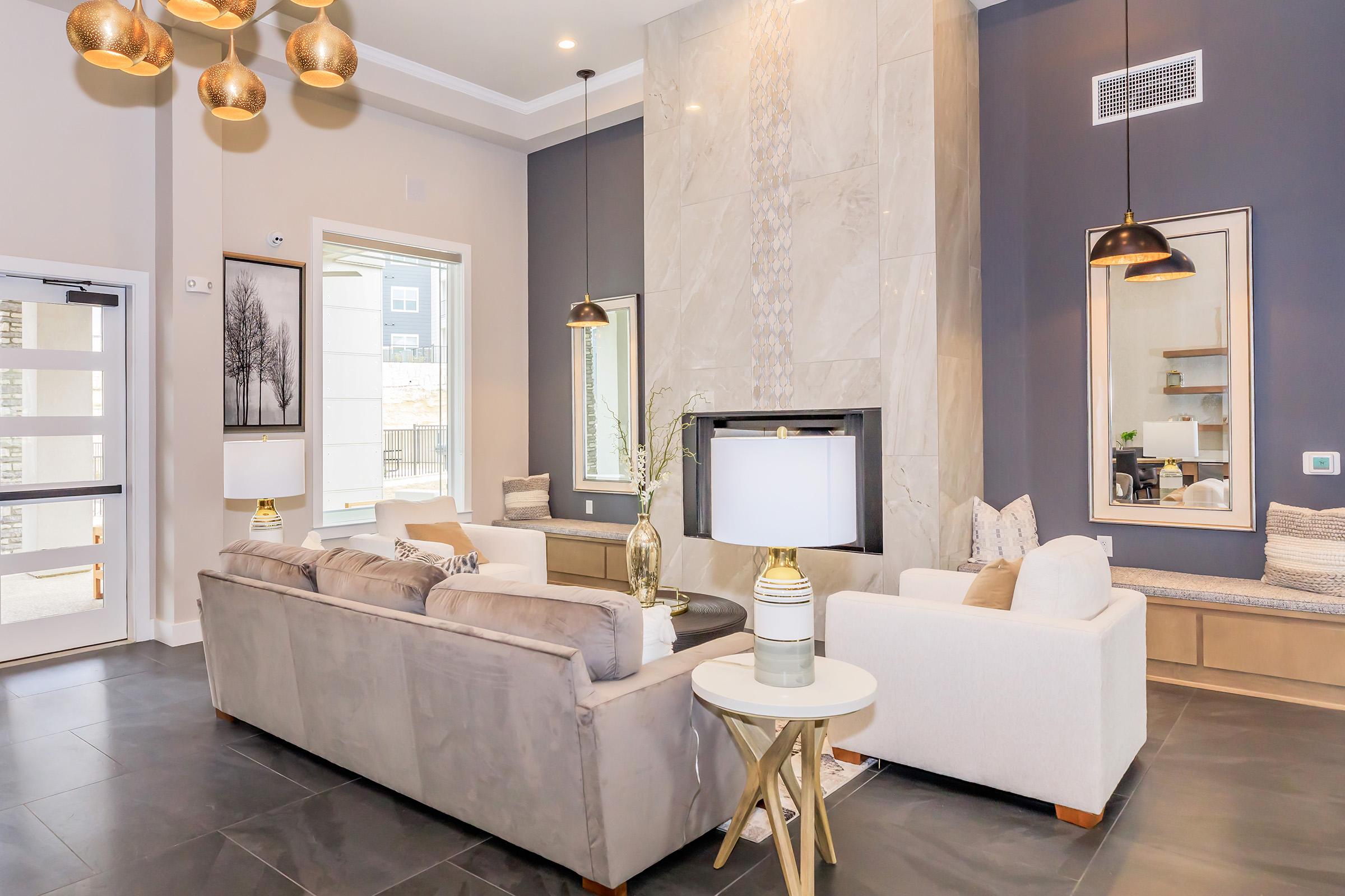 Modern living room featuring two sofas, decorative lamps, and stylish pendant lights. A fireplace is set against a marble accent wall, with large windows allowing natural light. A cozy seating area with decorative pillows and a coffee table enhances the inviting atmosphere.