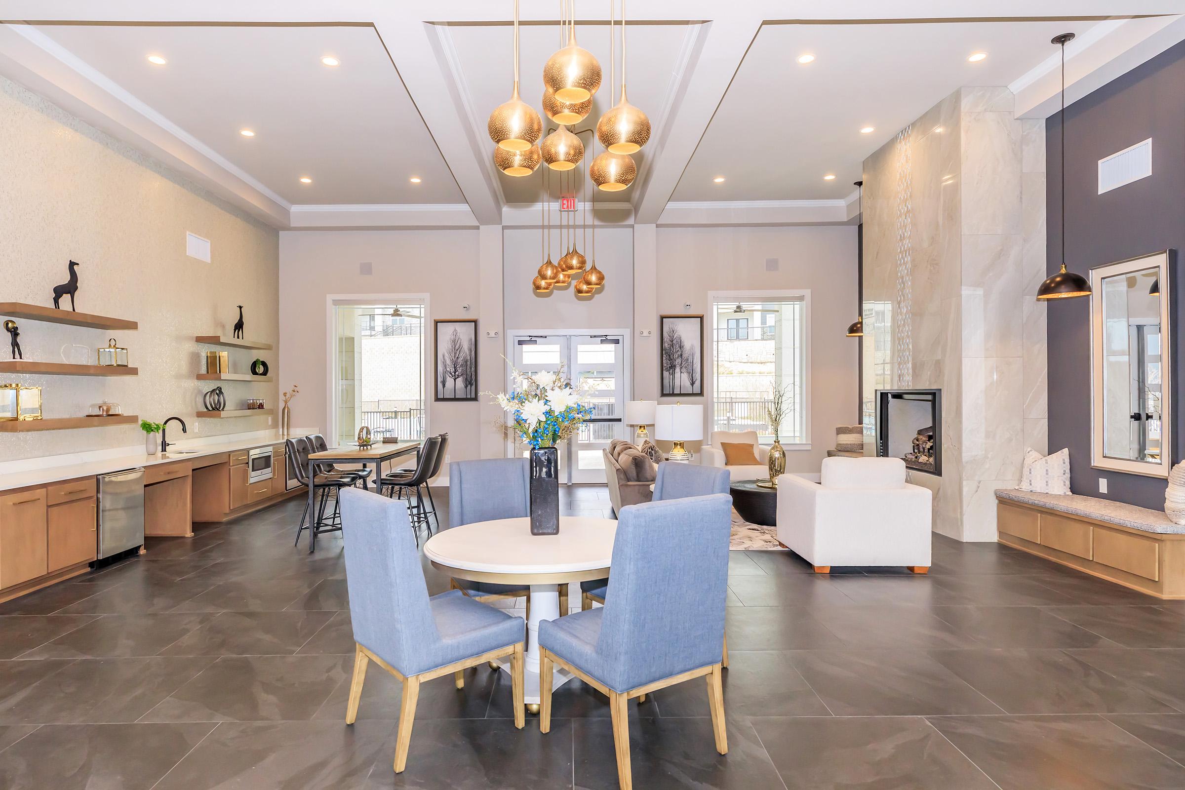 A modern, elegantly designed communal space featuring a round dining table with blue chairs, a stylish kitchen area, and a cozy seating arrangement. The room is enhanced by decorative lighting fixtures and a neutral color palette, with large windows allowing natural light and decorative accents throughout.