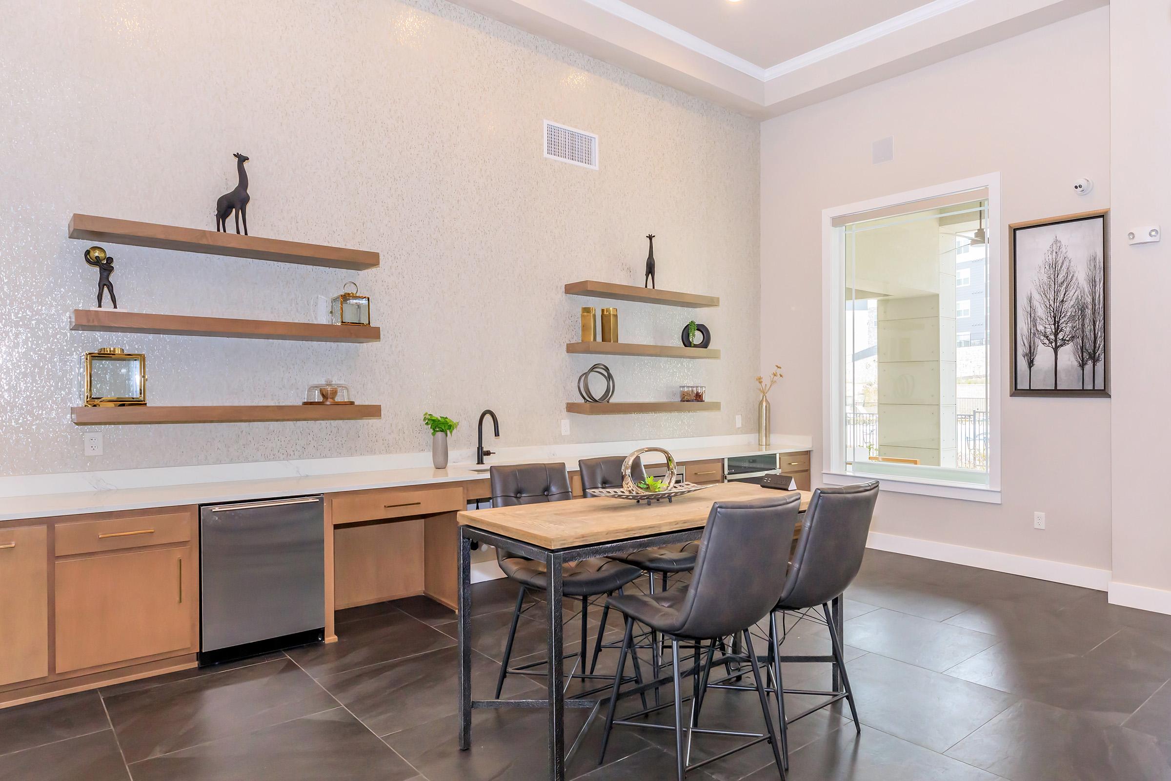 Modern kitchen and dining area with a large wooden table and black chairs. Open shelving on the walls holds various decorative items. Stainless steel appliances are visible, and large windows provide natural light. Neutral colors and sleek design elements create an inviting atmosphere.