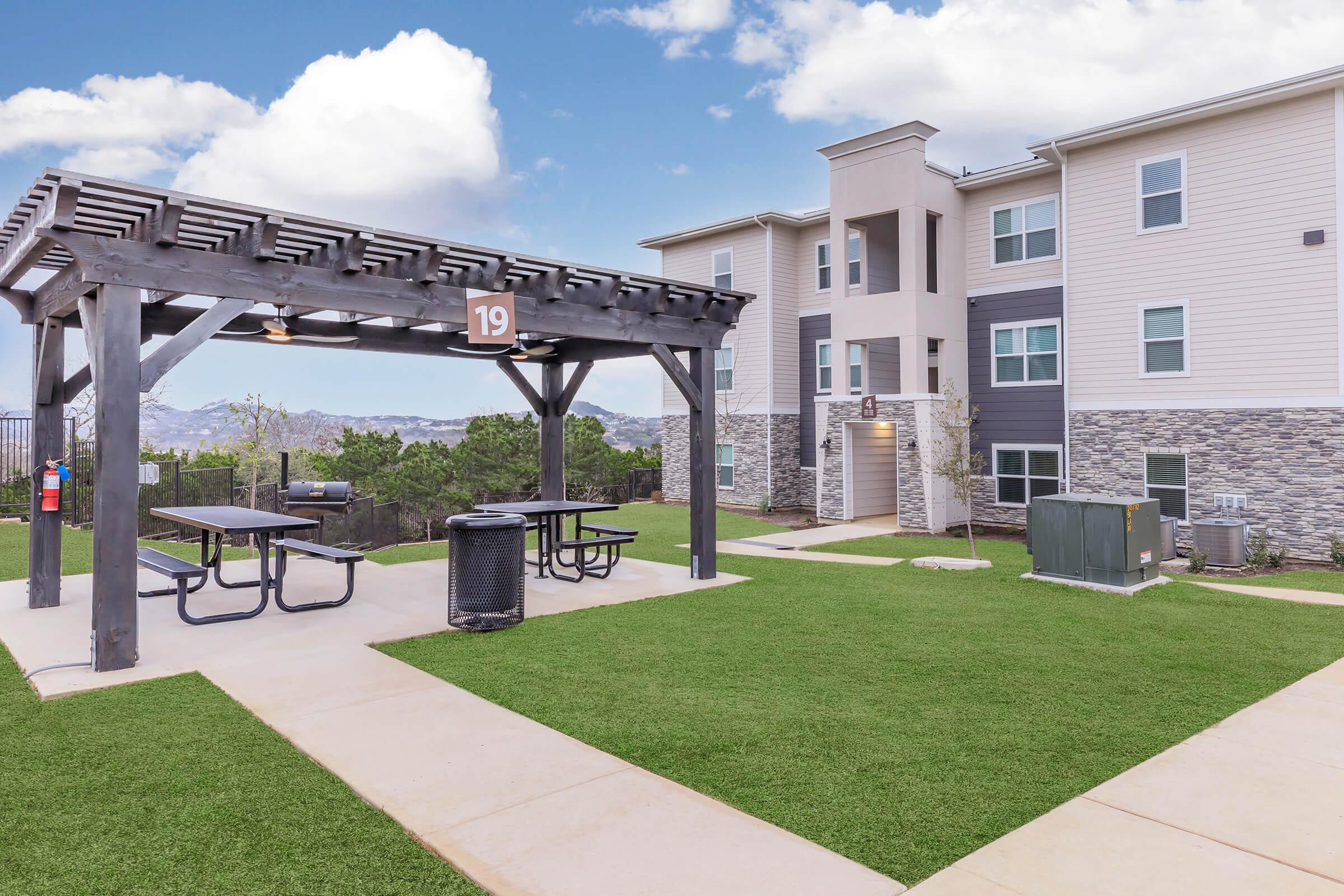 A landscaped outdoor area featuring a pergola with picnic tables, a grassy lawn, and a multi-story residential building in the background. The setting includes trash bins, landscaped greenery, and mountain views under a partly cloudy sky.
