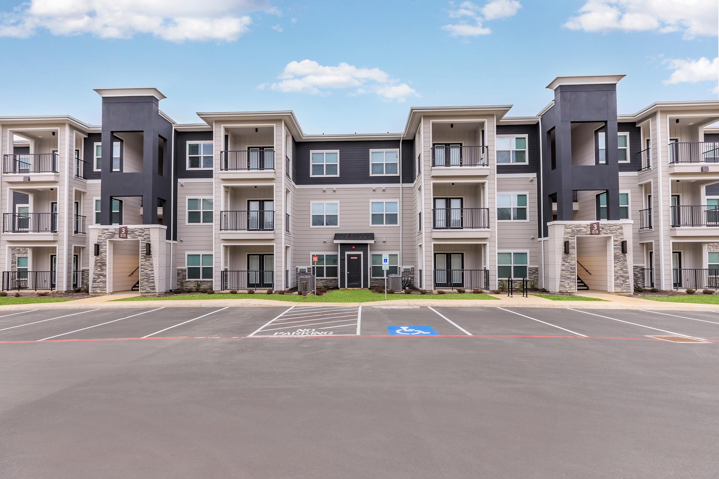 Modern apartment complex featuring multiple floors with balconies. The building has a combination of gray and beige siding, large windows, and a well-maintained parking lot with designated spaces. The sky is clear, indicating a bright day.
