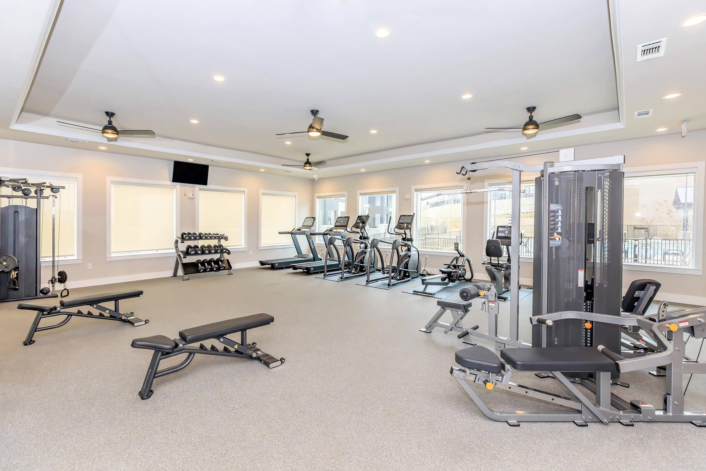 A bright, modern gym featuring various exercise equipment including treadmills, an elliptical machine, a weight bench, and a multi-station strength machine. Large windows allow natural light to flood the space, and there are ceiling fans for ventilation. The flooring is grey and well-maintained.