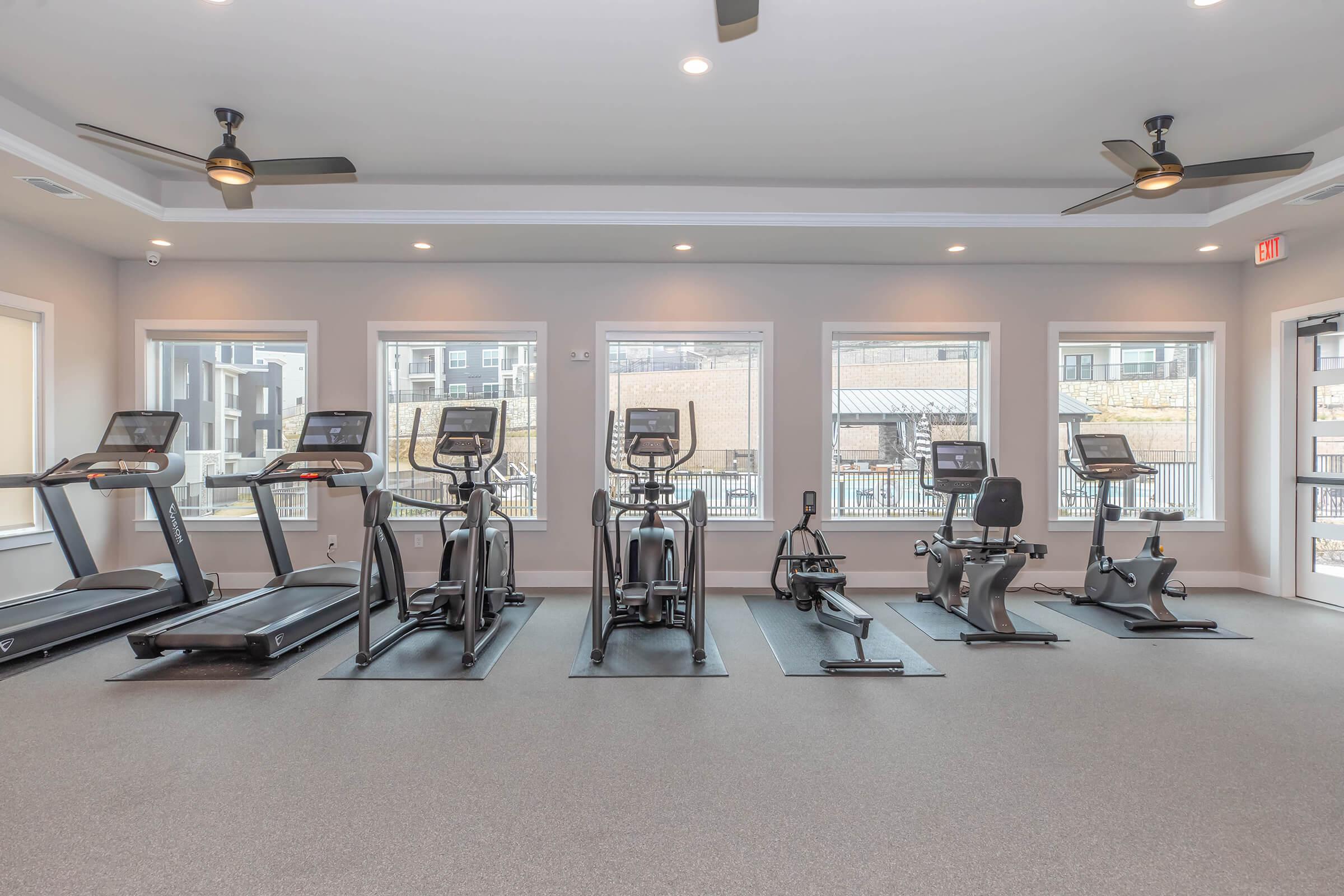 A modern gym featuring various cardio machines, including treadmills, ellipticals, and a stationary bike. The space is well-lit with large windows and has a clean, minimalistic design. Ceiling fans provide ventilation, enhancing the workout environment.