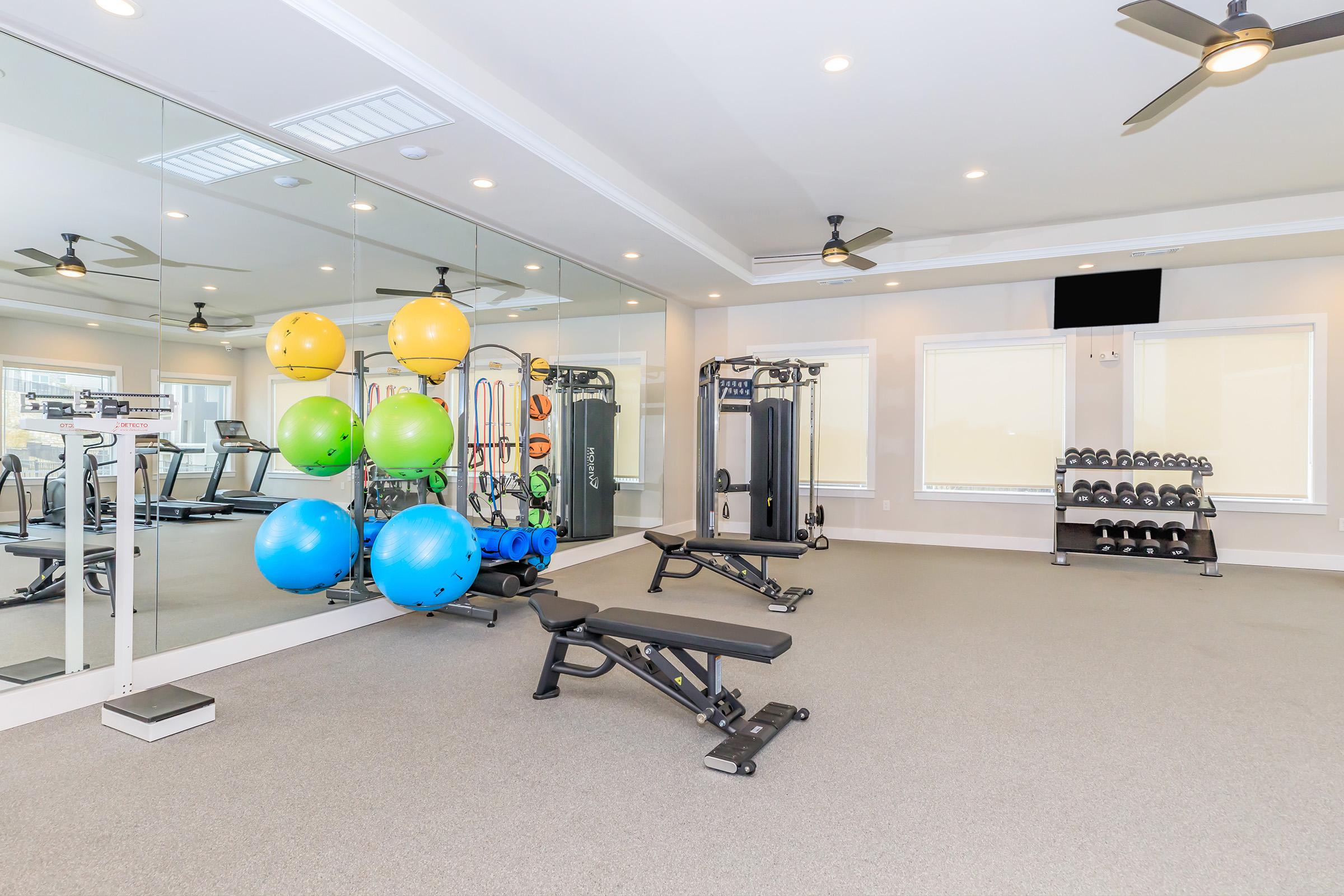 A modern gym interior featuring exercise equipment, including a weight bench, dumbbells, and a cable machine. Brightly colored stability balls are displayed against a mirror wall. The space is well-lit with natural light and ceiling fans, creating an inviting atmosphere for workouts.