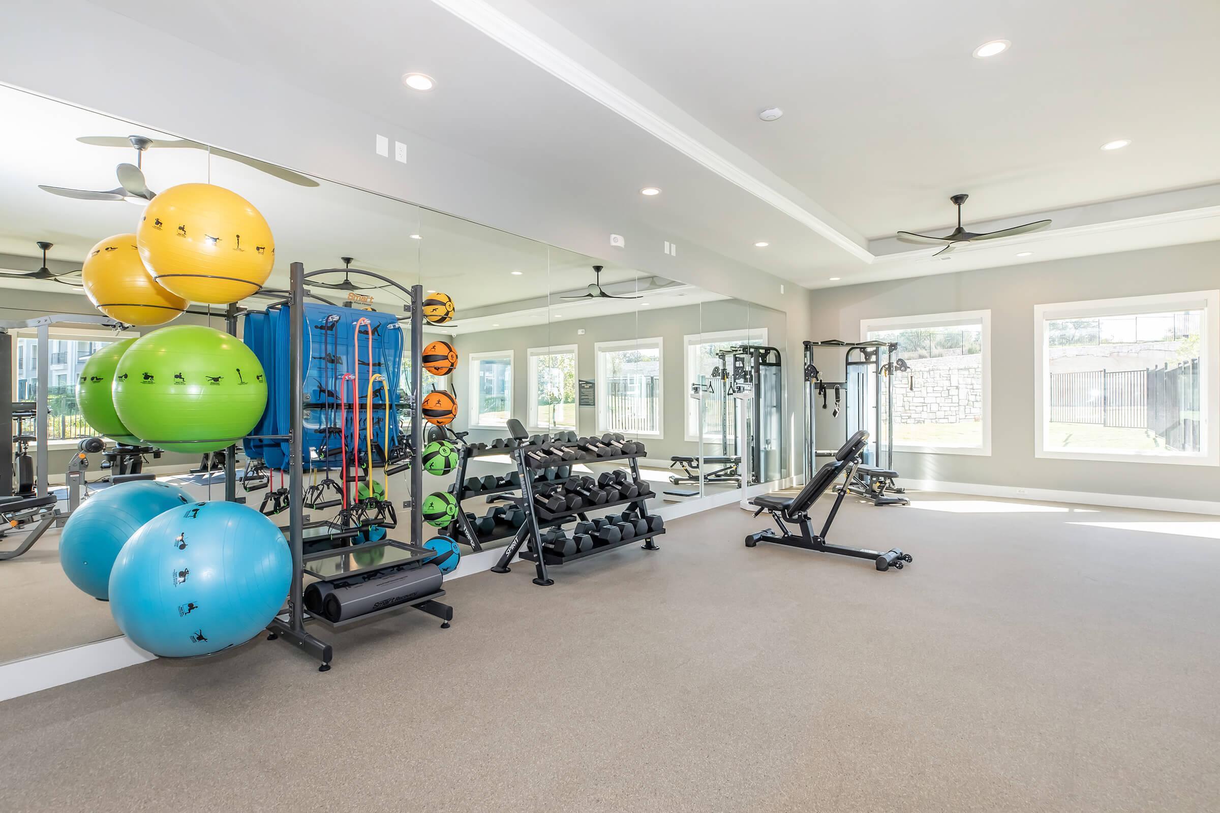 A modern gym with exercise equipment including dumbbells, resistance bands, and fitness balls in vibrant colors. The space features large windows allowing natural light, mirrors on the walls, and a clean, minimalist design. Ceiling fans provide ventilation for an inviting workout environment.