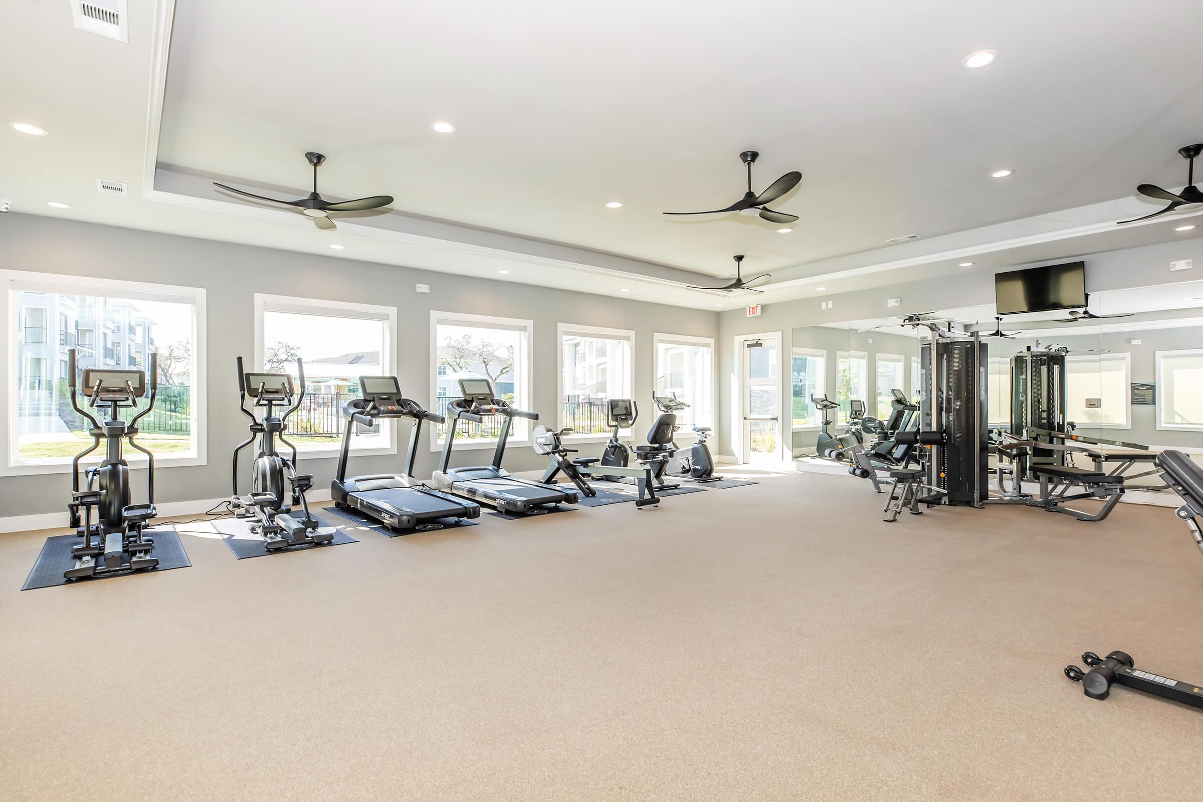 A modern gym featuring several exercise machines, including treadmills and stationary bikes, arranged neatly in a well-lit space. Large windows provide natural light, and there are fans on the ceiling for ventilation. Mirrors line one wall, enhancing the openness of the room.