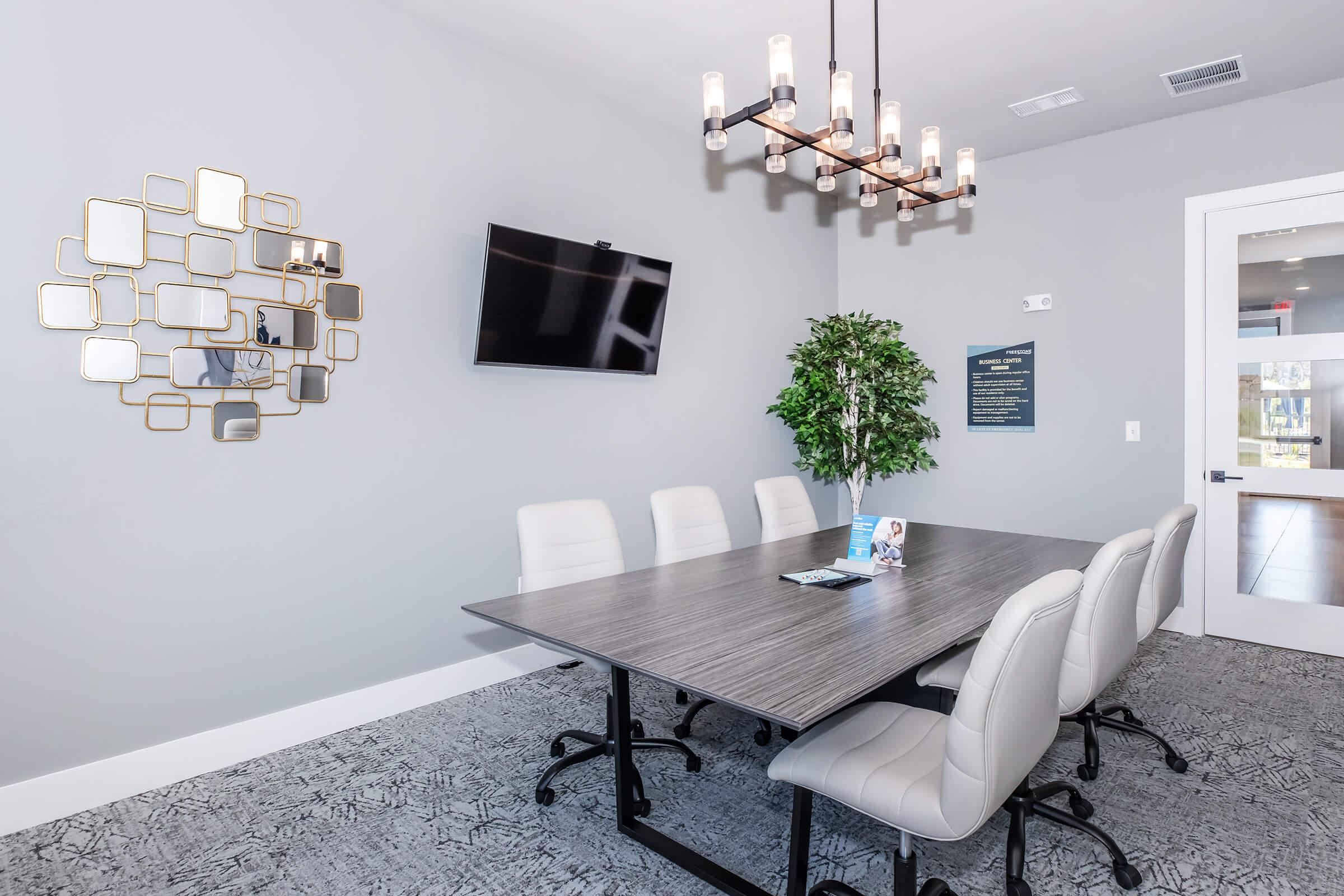 A modern conference room featuring a large table surrounded by six white chairs, a decorative wall mirror, a flat-screen TV mounted on the wall, and a potted plant. The room is well-lit by a stylish chandelier and has light gray walls and a textured gray floor.