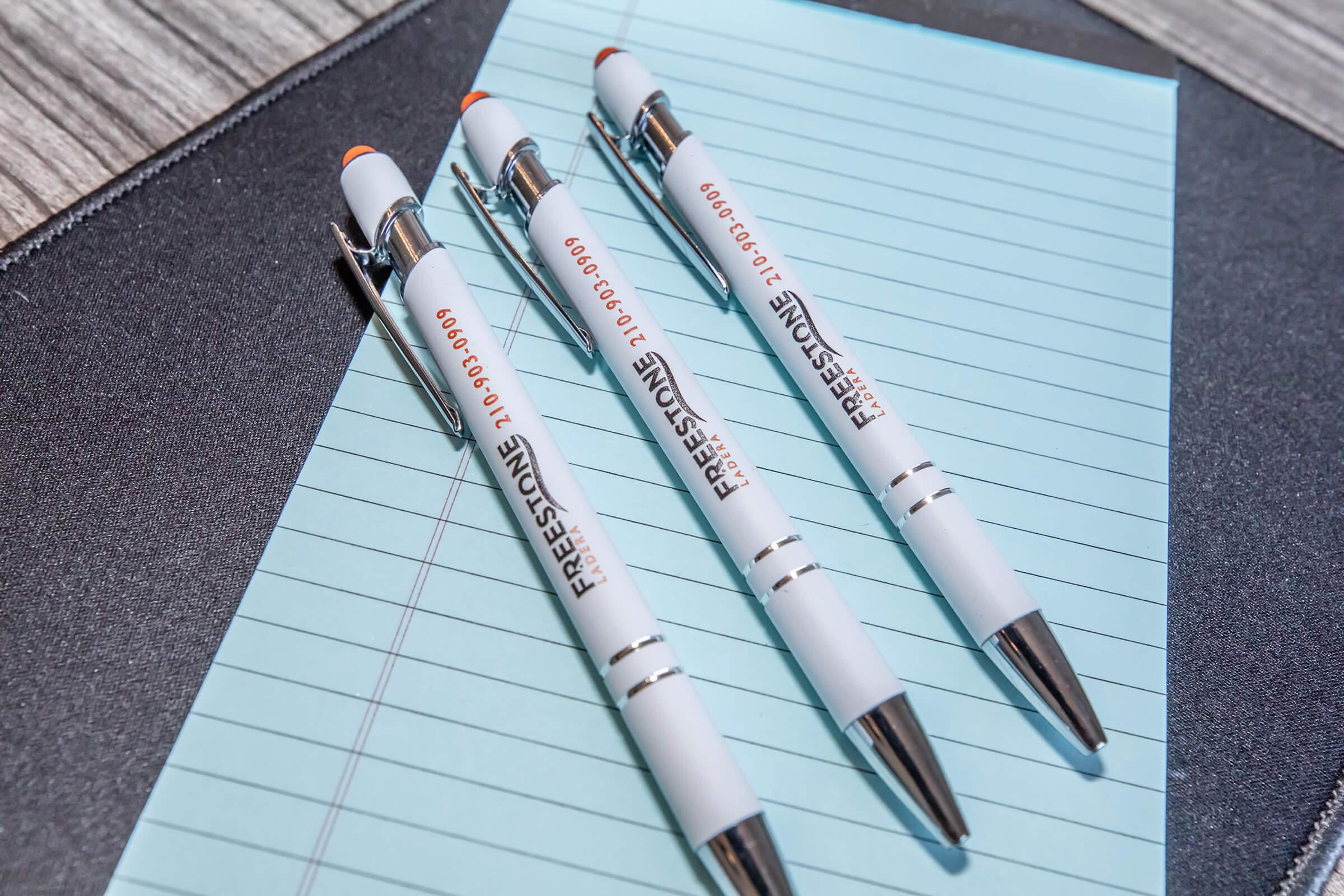 Three white ballpoint pens with black ink resting on a light blue notepad. The pens are imprinted with the word "FREESTONE" and a phone number. The notepad features lined pages and is placed on a dark textured surface.