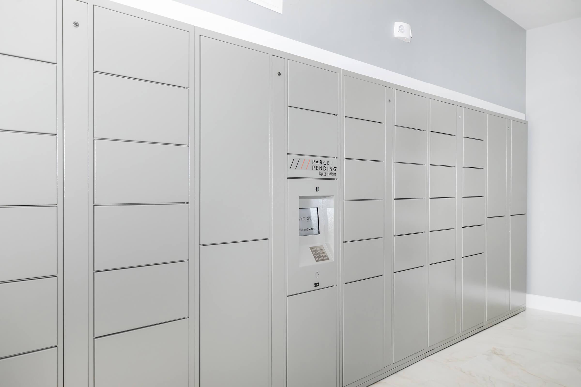 A row of gray parcel lockers mounted against a light gray wall. The lockers are designed for package delivery and retrieval, featuring a central kiosk with a display and keypad for user interaction. The space is well-lit and modern, with a clean, minimalist aesthetic.