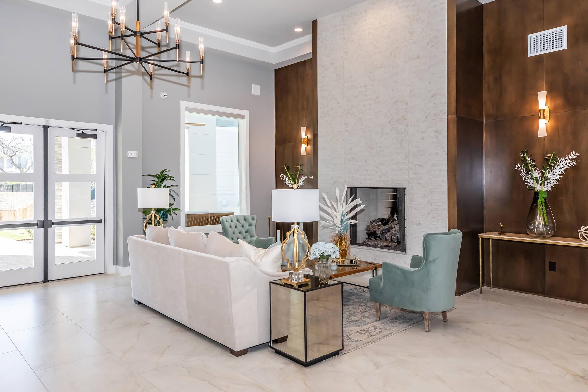 A modern, stylish living room featuring a light-colored sofa, a green armchair, and a decorative coffee table. The decor includes a chandelier, potted plants, and a stone fireplace. Large windows let in natural light, enhancing the elegant atmosphere with neutral tones and contemporary furnishings.