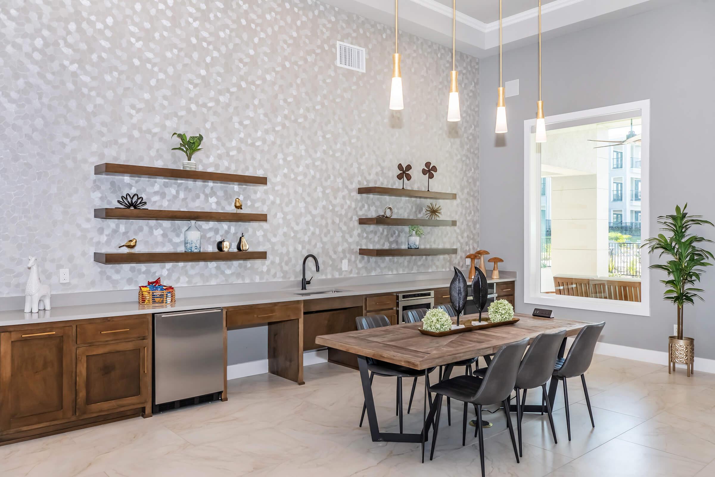 A modern kitchen and dining area featuring a wooden dining table with black chairs, light wood cabinets, and decorative shelving against a white speckled wall. The space includes pendant lighting, a silver refrigerator, and a sink, with plants and decorative items on the shelves. A large window allows natural light to enter.