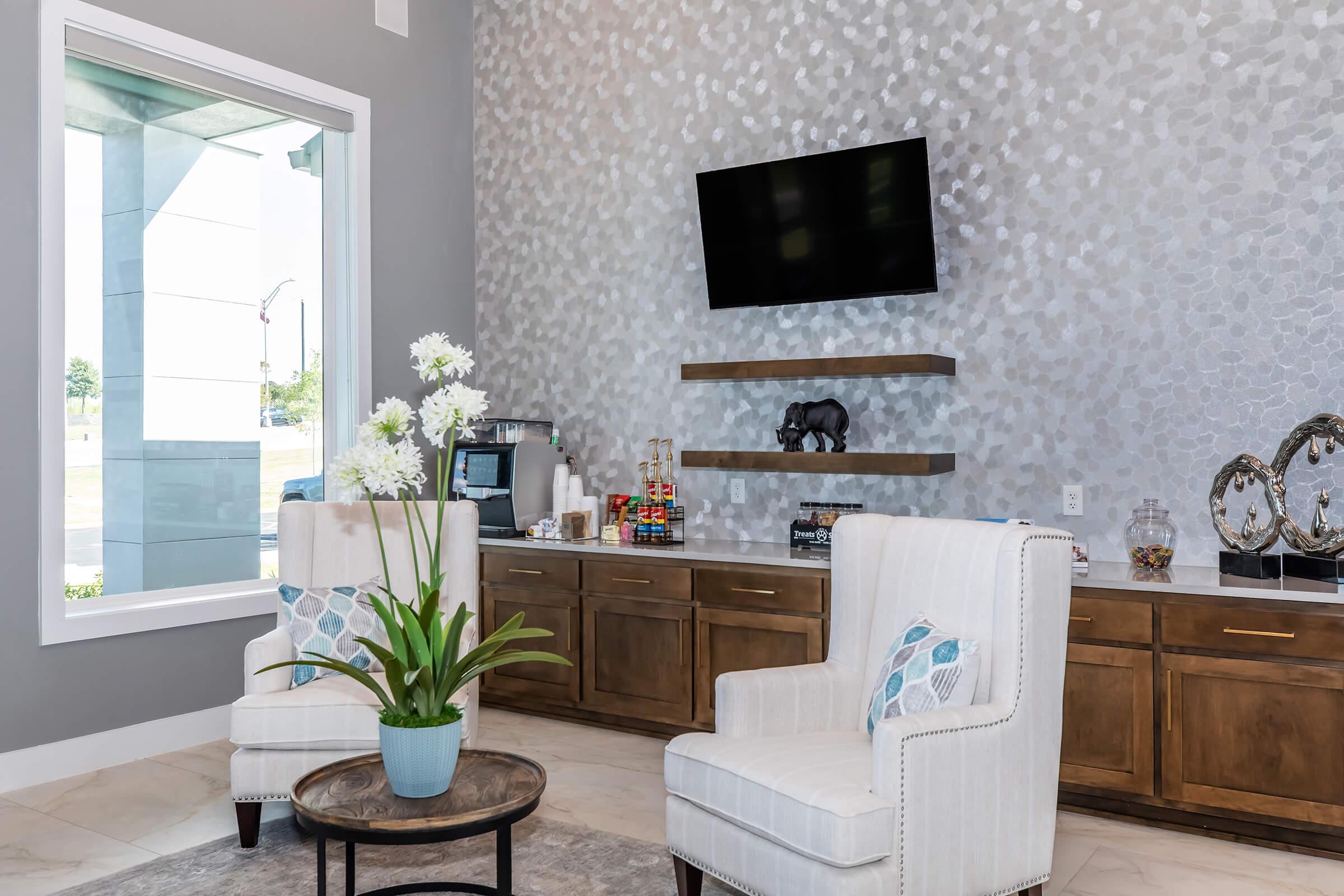 A modern reception area featuring two white upholstered chairs, a round coffee table with a potted plant, and a wooden cabinet displaying snacks and decorative items. A flat-screen TV is mounted on a stylish wall with a textured gray finish, creating a welcoming atmosphere.