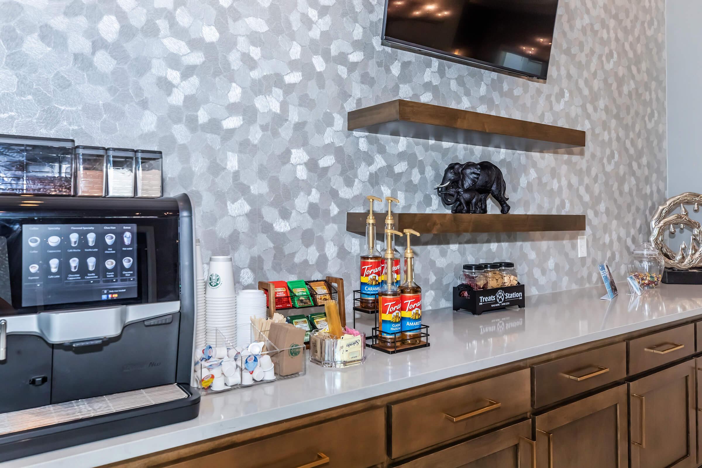 A modern coffee station featuring an espresso machine, condiments, and various coffee-related items. There are decorative shelves with a small elephant sculpture and a decorative piece. The backdrop features a textured wall design, and a flat-screen TV is mounted above.