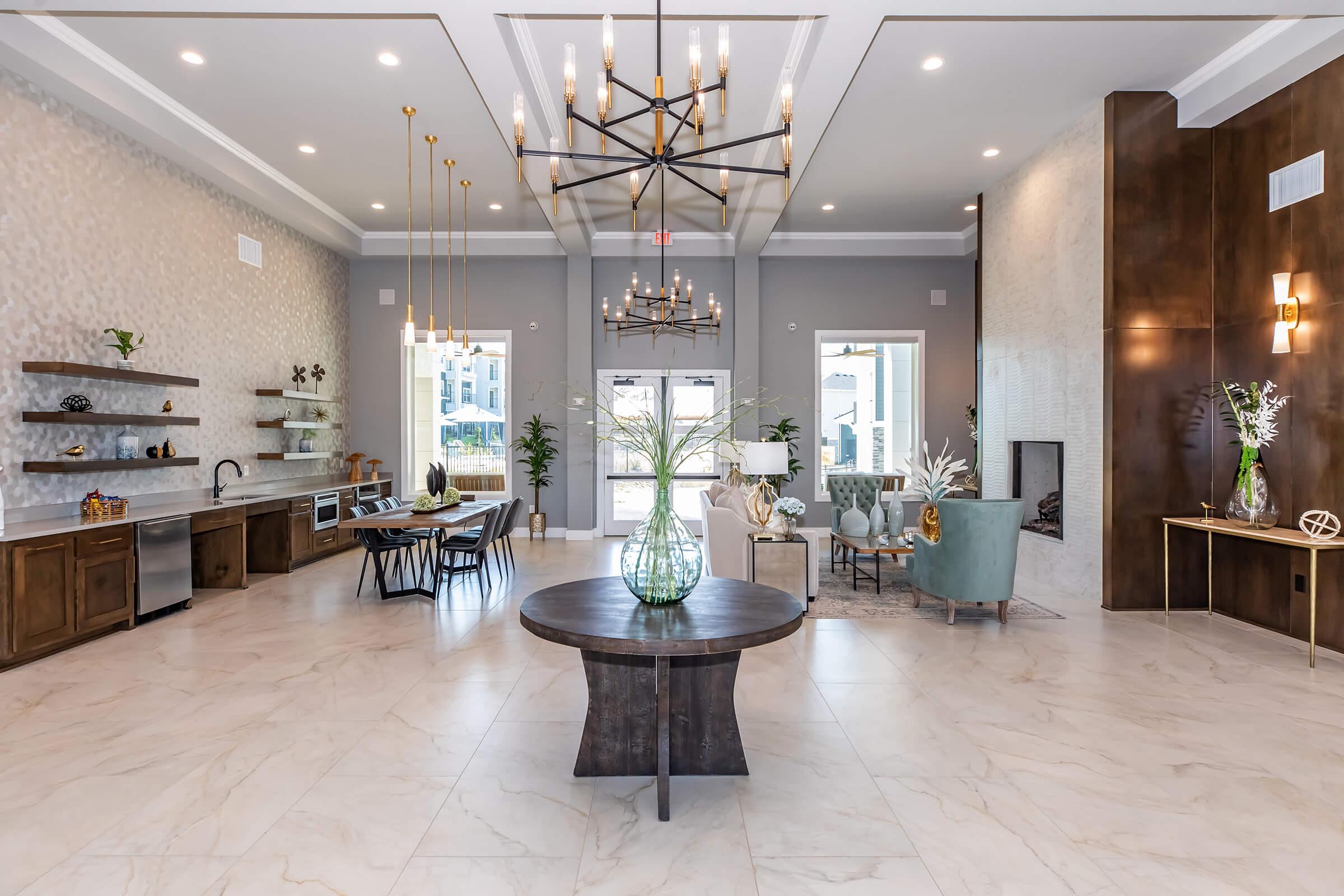 A spacious and modern lobby featuring elegant furnishings. The area includes a circular wooden table, a green upholstered chair, a dining area with a wooden table and black chairs, and decorative plants. Large windows allow natural light to fill the space, which is styled with contemporary light fixtures and artwork on the walls.