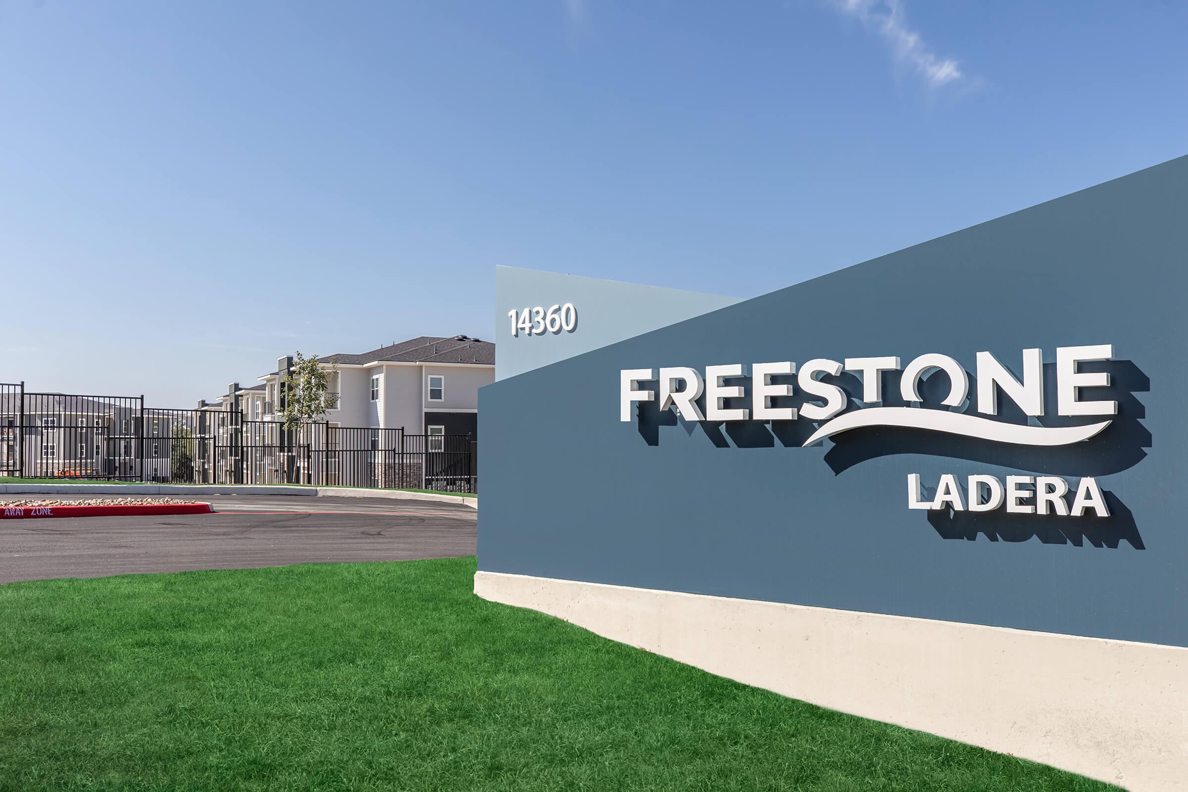 Sign for Freestone Ladera, featuring the name prominently displayed in white letters on a blue background. The entrance to an apartment complex is visible in the background, with modern buildings and a well-maintained green lawn. Clear blue sky above.