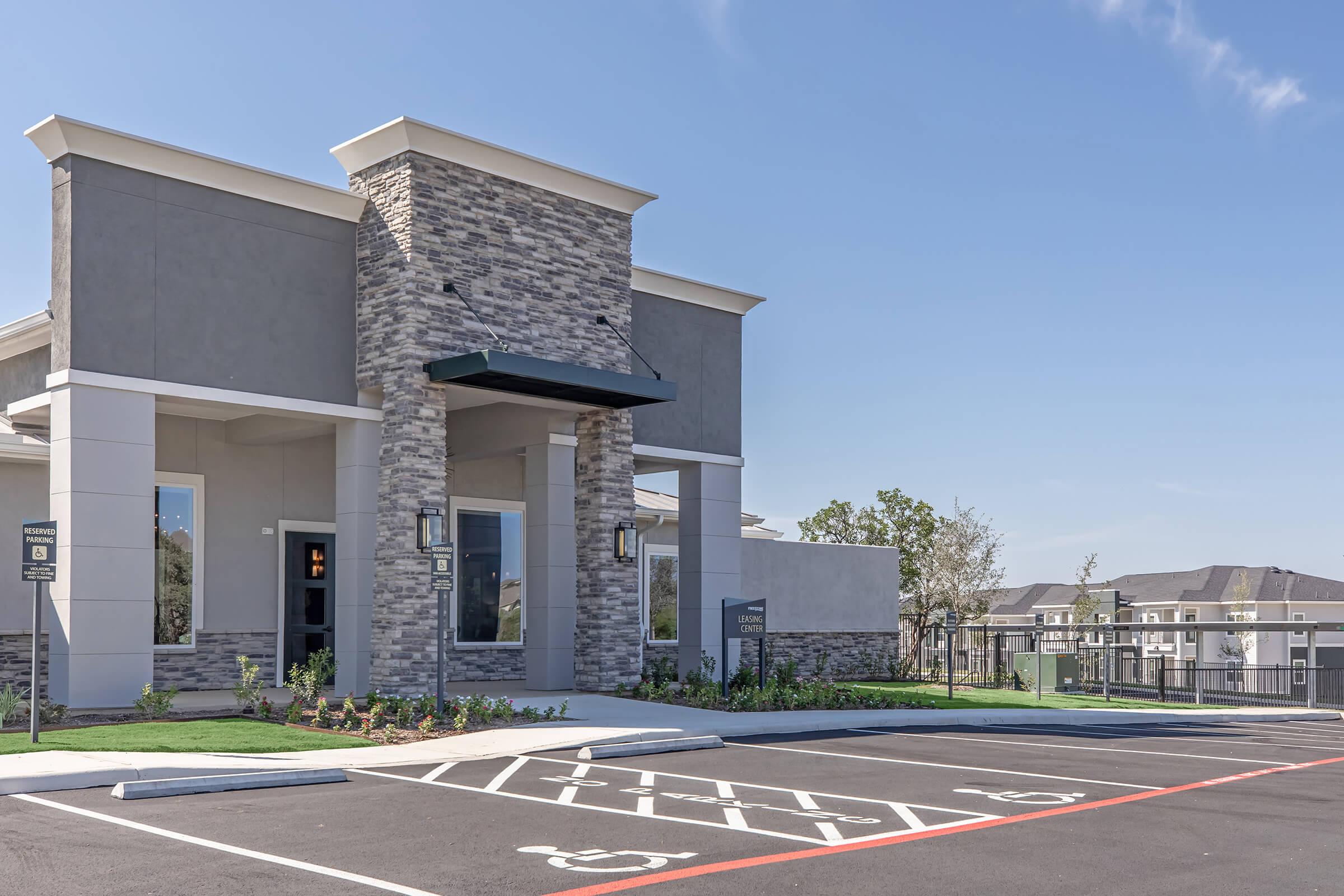 Modern building exterior featuring a stone-accented entrance, large windows, and an overhang. The surrounding area has landscaped greenery, a clearly marked parking lot with reserved spaces. In the background, additional residential buildings are visible under a clear blue sky.