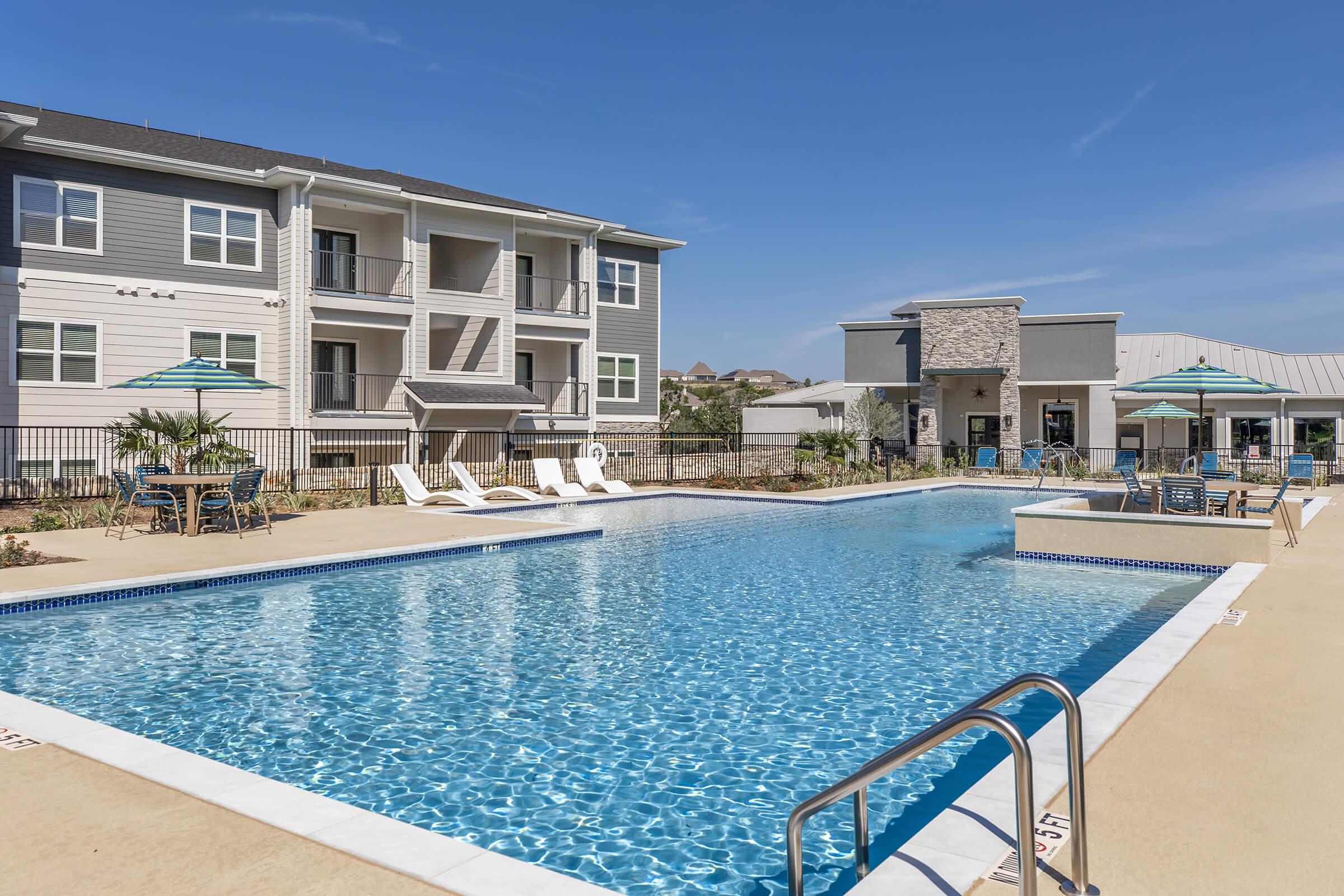 A sparkling outdoor swimming pool surrounded by lounge chairs and umbrellas, with an apartment building in the background. The setting features well-maintained landscaping and a clear blue sky, creating a welcoming atmosphere for relaxation and leisure.