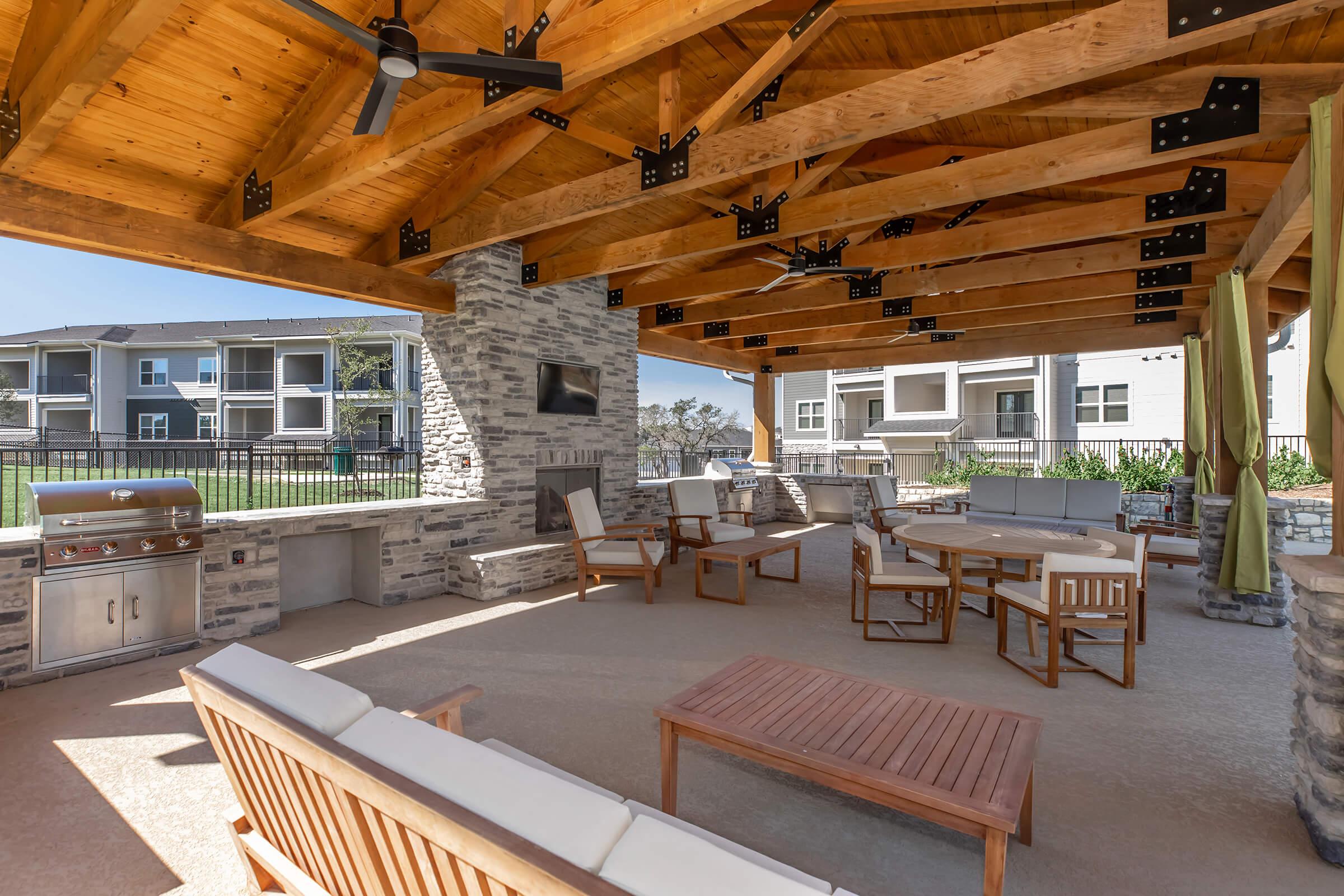 An outdoor patio area featuring a wooden ceiling with exposed beams, stone accents, and a grill. The space includes comfortable seating with a wooden table and chairs, perfect for relaxation or entertaining, and is surrounded by lush greenery and modern apartment buildings in the background.