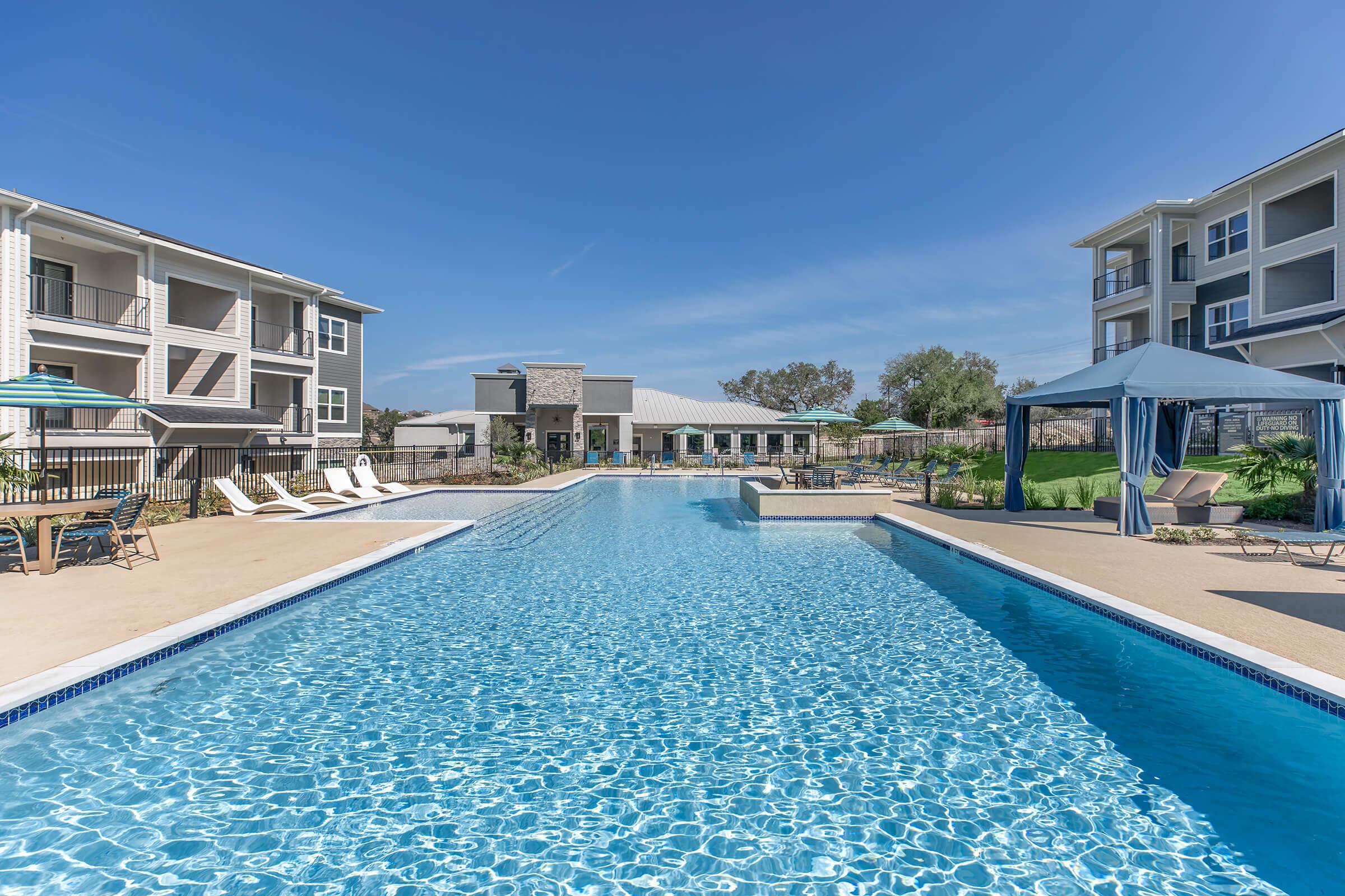 A bright and inviting outdoor swimming pool surrounded by lounge chairs and umbrellas. Apartment buildings are visible in the background, along with a well-maintained patio area. Clear blue skies enhance the relaxing atmosphere of the leisure space.