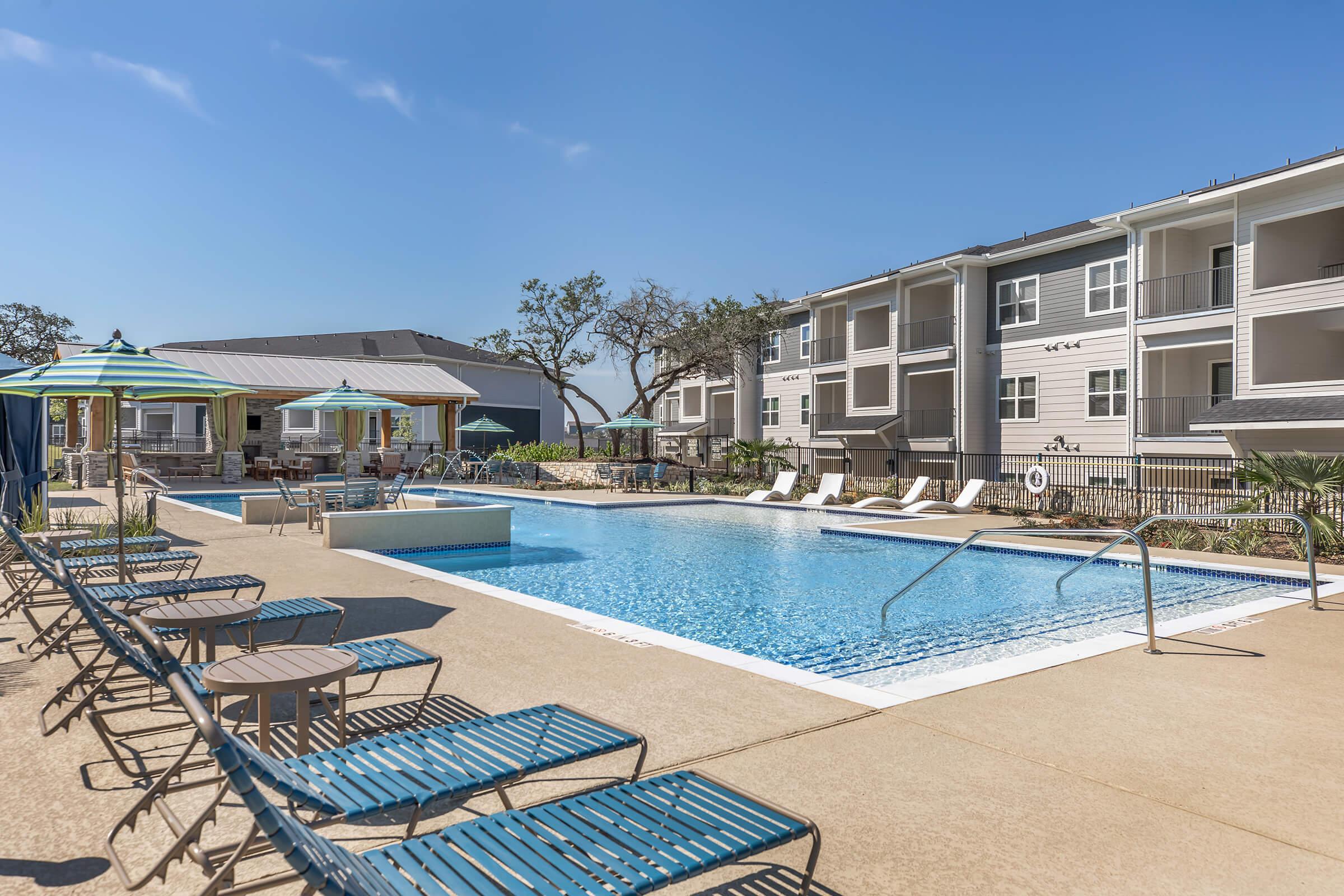 A bright swimming pool area surrounded by lounge chairs, umbrellas, and greenery. The pool is clear and inviting, with a shallow section and nearby seating. Modern apartment buildings are visible in the background under a clear blue sky.