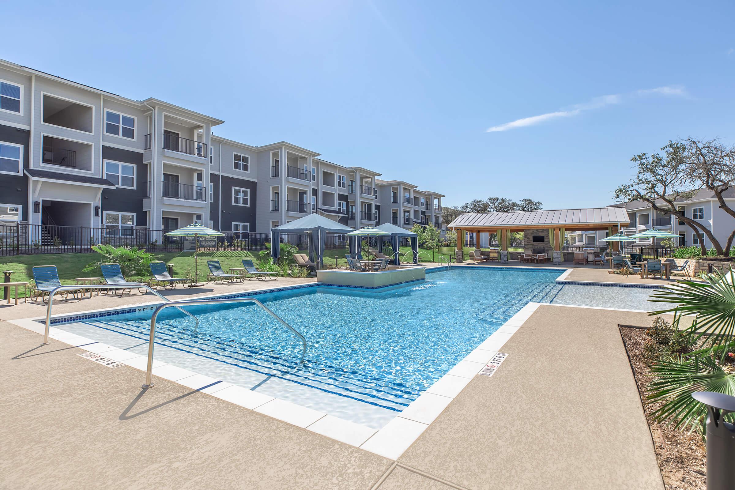 Swimming pool with clear blue water, surrounded by lounge chairs and umbrellas. In the background, there are modern multi-story residential buildings. A shaded gazebo is visible to the right, creating a relaxing outdoor space. Bright sunny sky with a few clouds overhead.