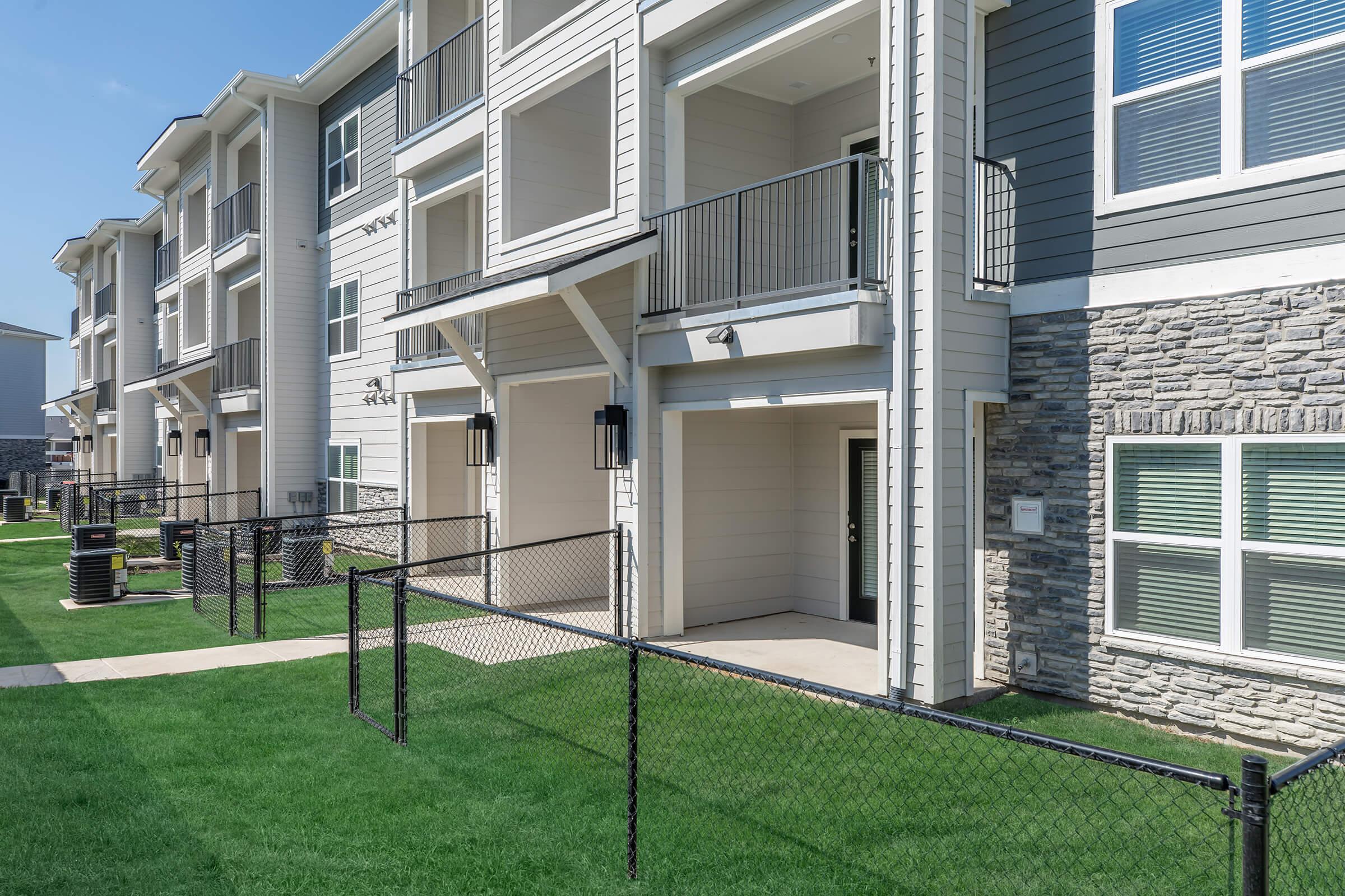 Modern apartment complex featuring multiple buildings with balconies, large windows, and a stone accent. A manicured lawn is visible in front, bordered by chain-link fencing. The sky is clear and blue, creating a bright atmosphere.
