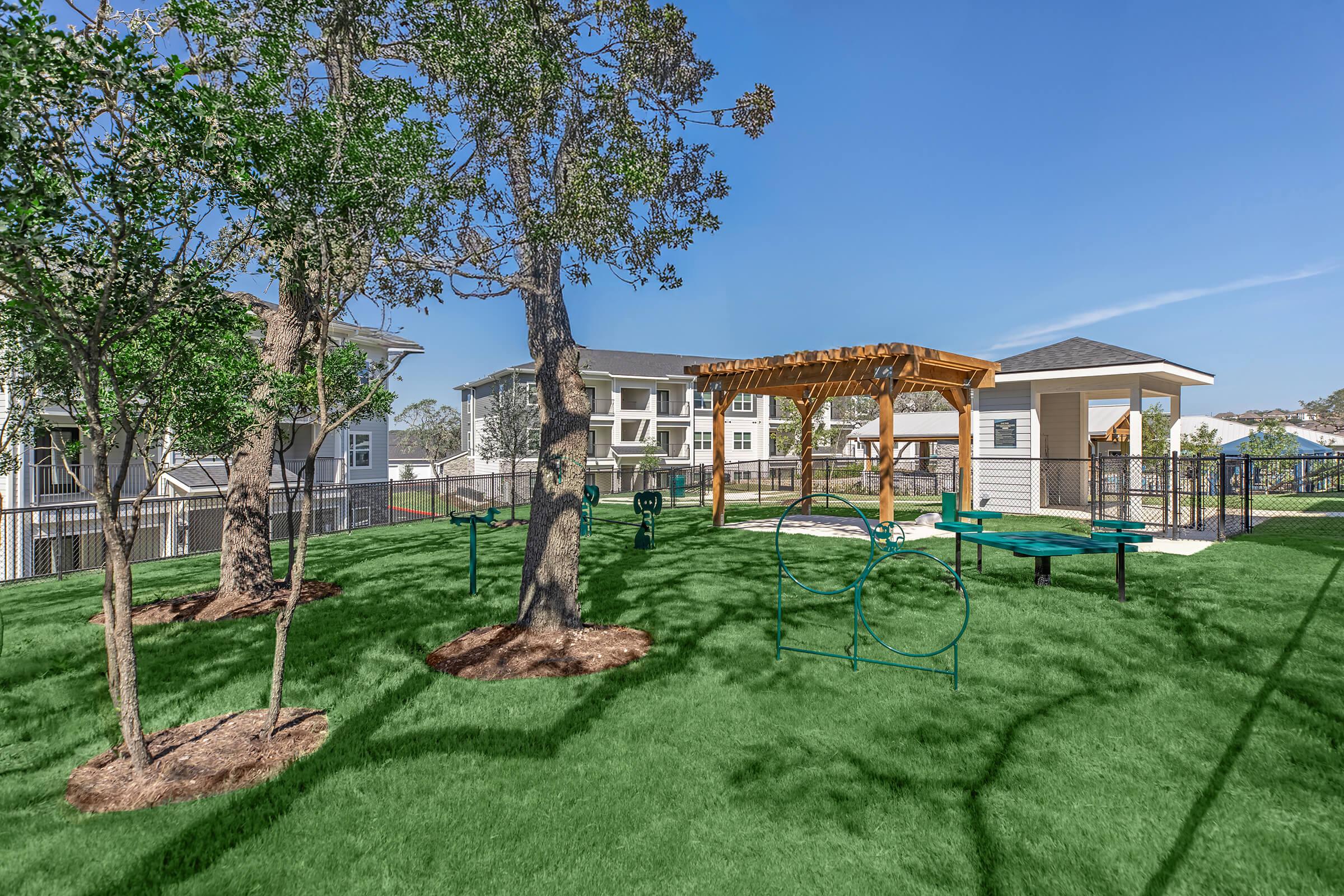 A well-maintained outdoor play area featuring green grass, trees, and a wooden pavilion. The space includes playground equipment and picnic tables, surrounded by a fence. Bright blue sky and residential buildings in the background create a welcoming atmosphere for community gatherings and activities.