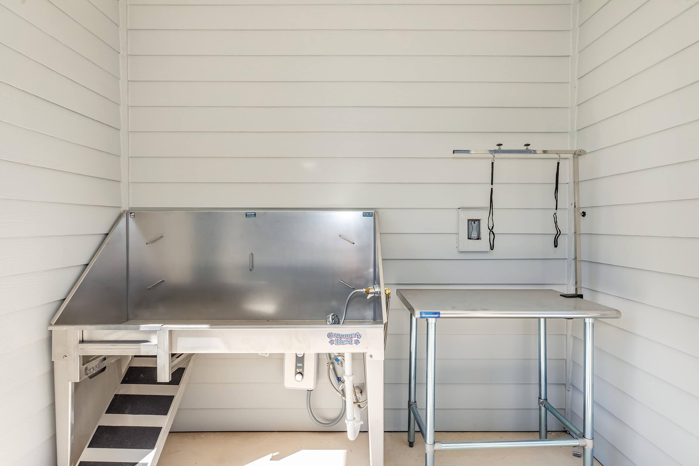 A clean, modern pet washing station featuring a stainless steel tub with an adjustable shower head, alongside a stainless steel table. The wall is made of white paneling, providing a bright and tidy appearance.