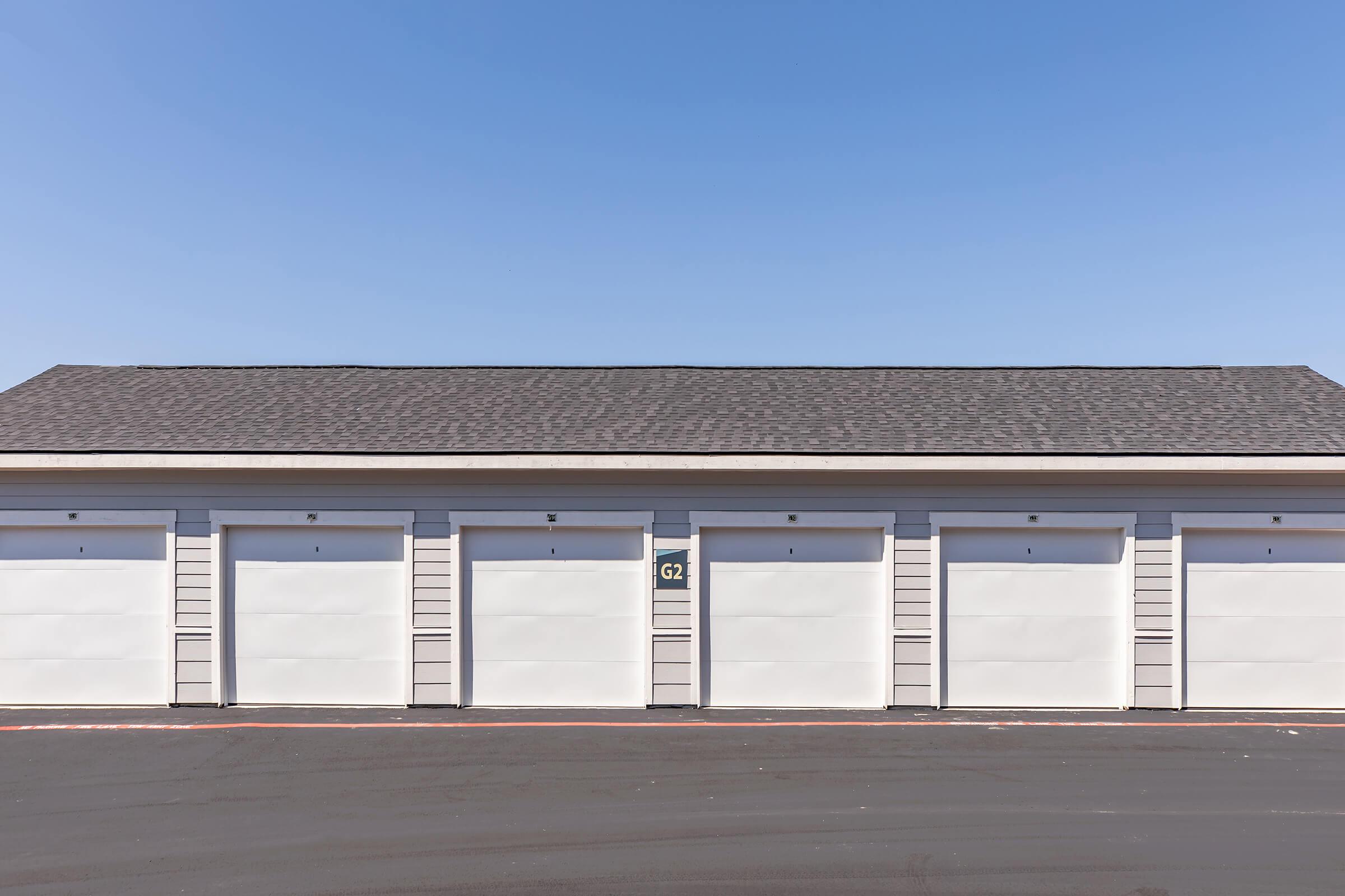 A row of five identical white garage doors with grey siding, all closed, under a clear blue sky. The garage doors are numbered, with one door marked "62." The asphalt driveway in front is clean and well-maintained. The scene conveys a neat and organized storage space.