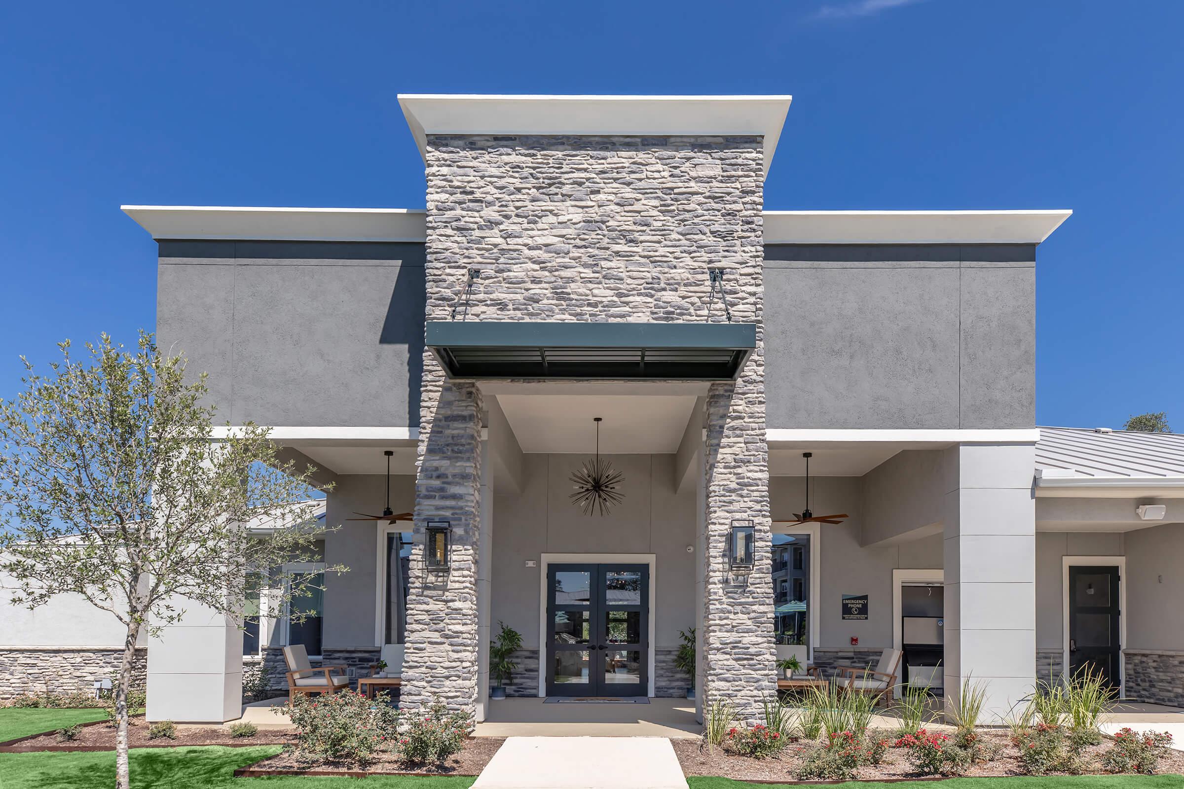Modern building facade featuring a combination of stone and stucco materials. The entrance is prominently displayed with a large, glass door flanked by windows. Decorative lighting fixtures hang on either side, and lush landscaping with young trees and shrubs is evident in the foreground against a clear blue sky.