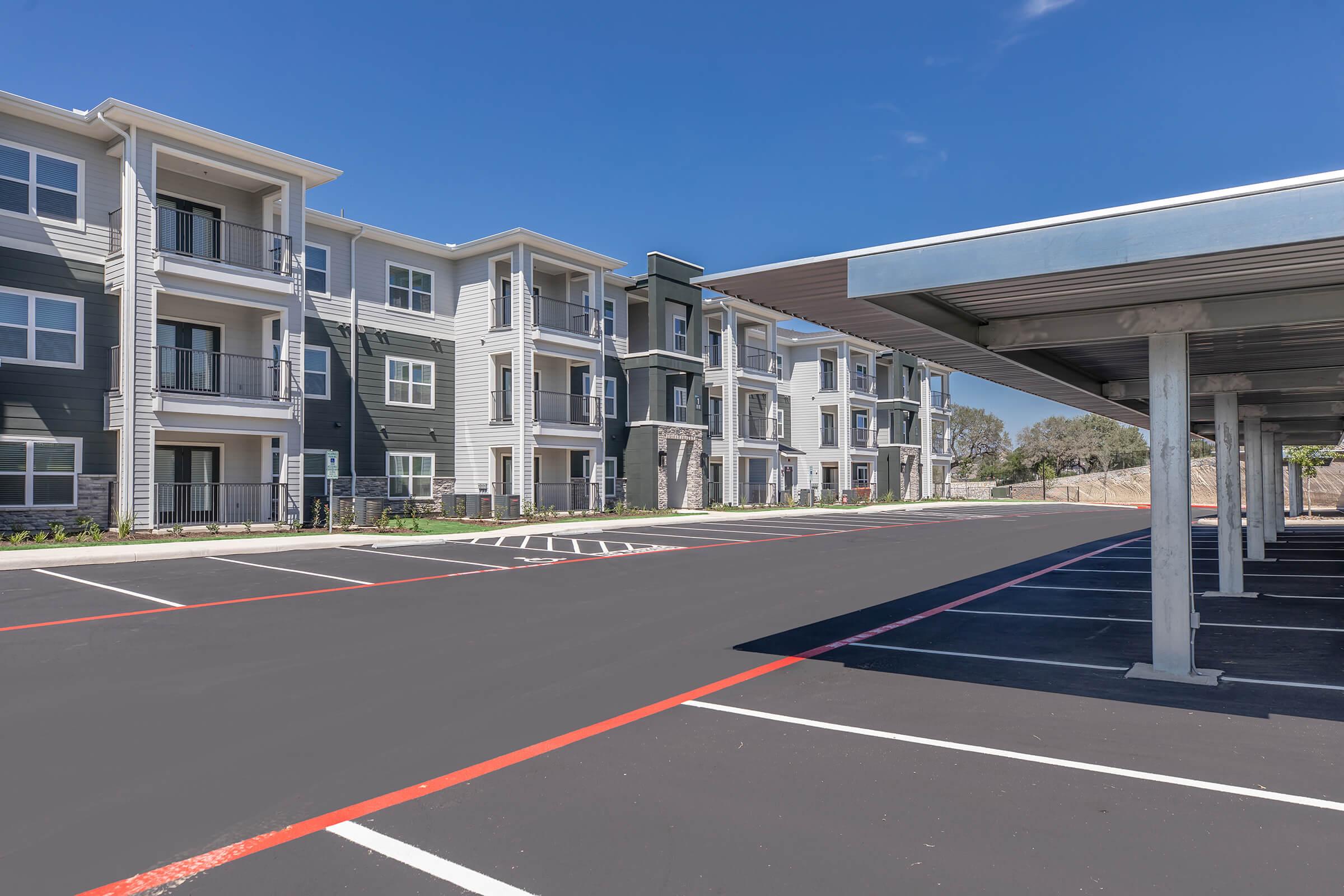 A modern apartment complex featuring multiple three-story buildings with balconies, surrounded by a parking lot. The parking lot has marked spaces and some covered parking areas. The sky is clear and blue, creating a bright and inviting atmosphere.