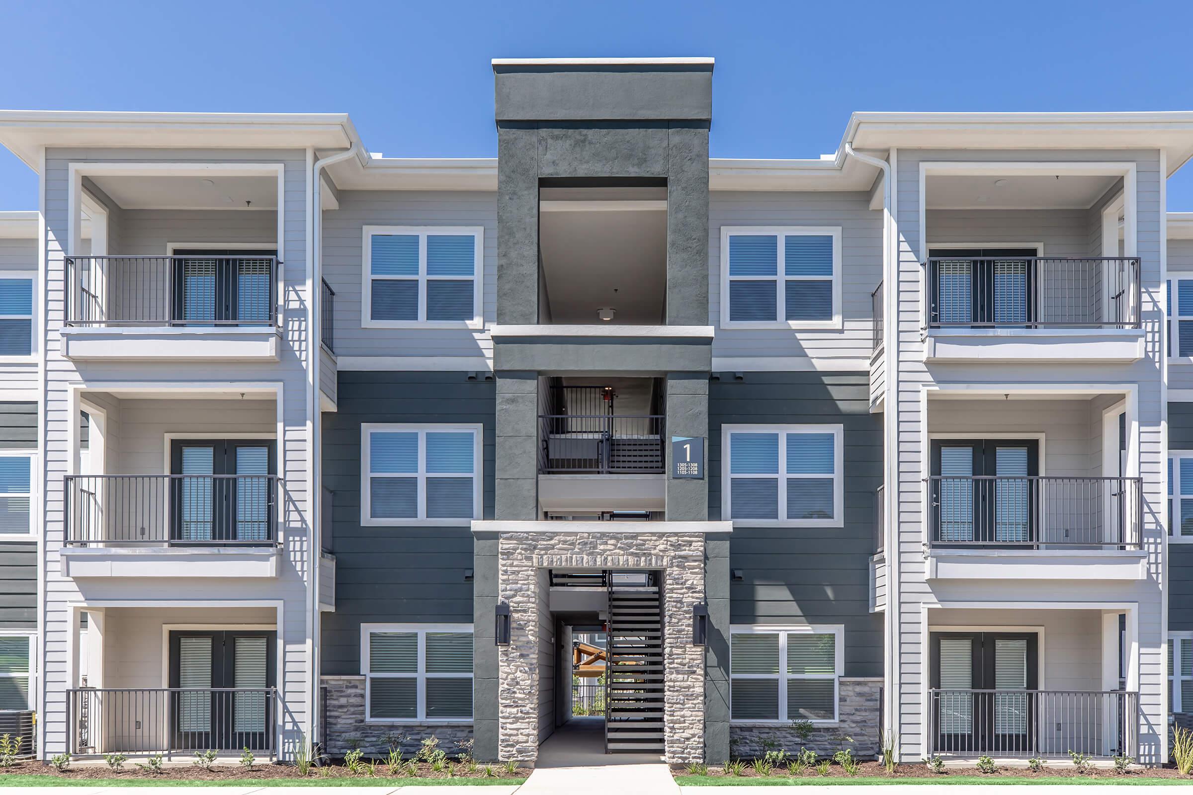 A modern multi-story apartment building featuring a mix of gray tones and stone accents. The façade includes several balconies, large windows, and a central entrance with stairs leading up. The surroundings are well-maintained with greenery visible at the base. Bright blue sky overhead.