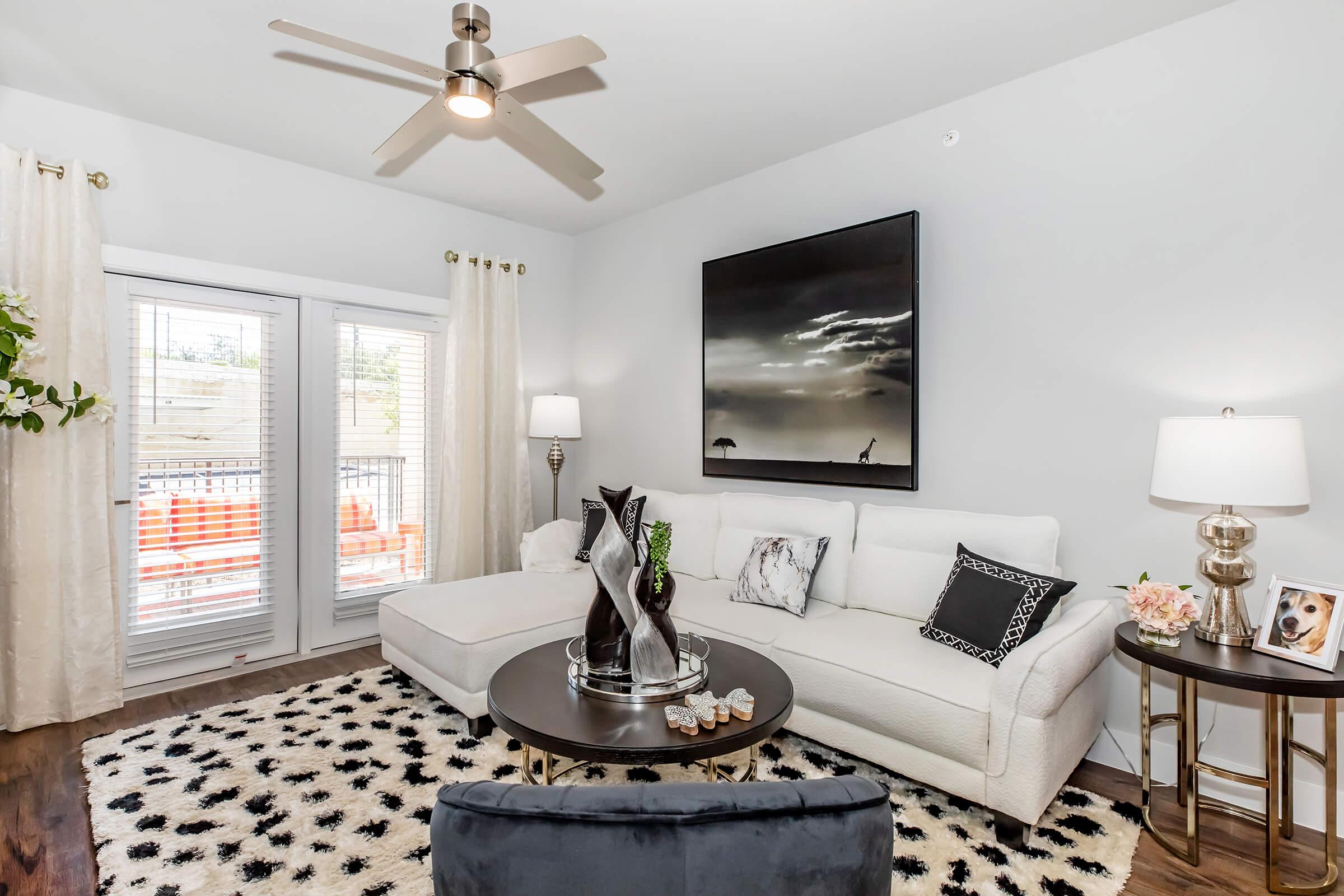 A modern living room featuring a white sectional sofa, a round coffee table adorned with decorative items, and a large wall-mounted artwork. Natural light streams through the windows dressed with light curtains, and there are potted plants and stylish lamps enhancing the decor.