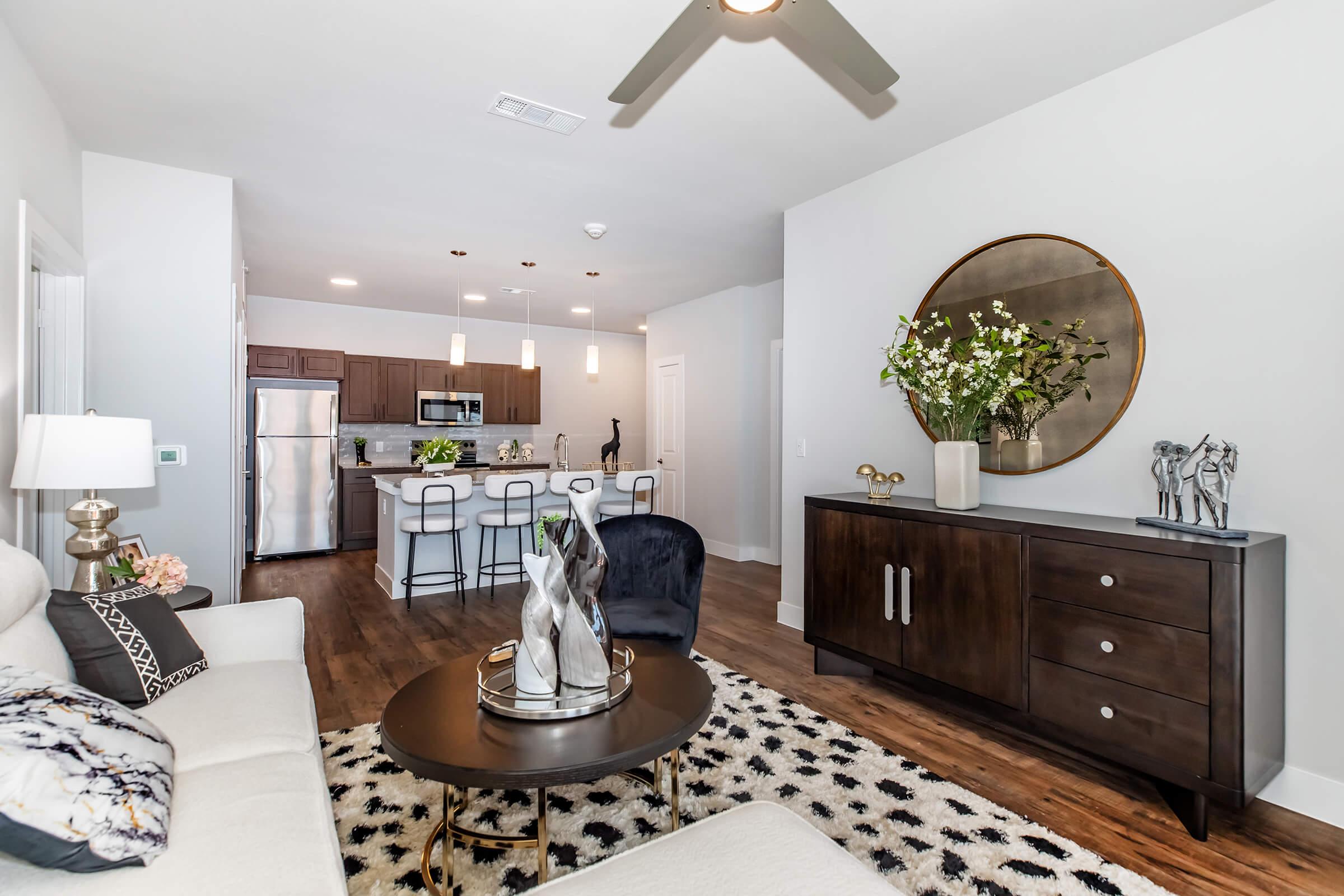 A stylish living room featuring a light-colored sofa, a round coffee table, and a decorative rug. In the background, there is a modern kitchen with dark cabinetry and stainless steel appliances. A large round mirror and vases with flowers are displayed on a dark wooden cabinet, creating an inviting atmosphere.