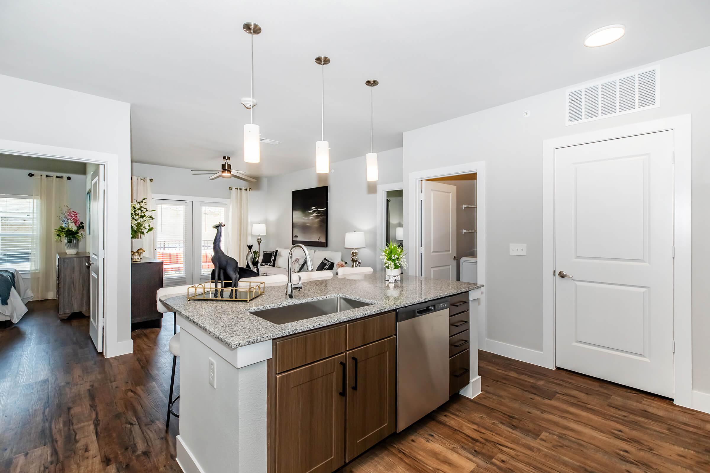 Modern kitchen featuring a granite island with a sink, stainless steel appliances including a dishwasher, and pendant lighting. The open layout connects to a living area with natural light from large windows, complemented by stylish decor and plants.