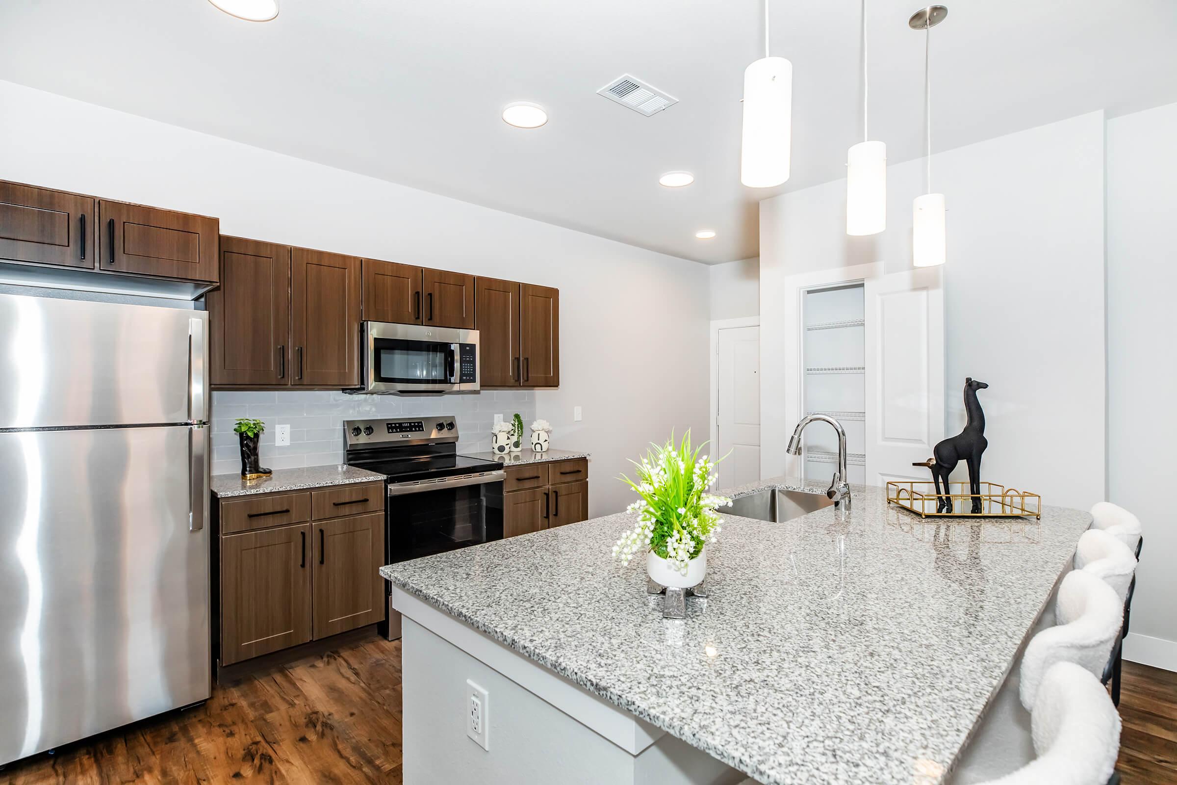 A modern kitchen featuring dark wood cabinetry, stainless steel appliances, and a large granite island with seating. The space is well-lit with pendant lighting and has a contemporary design, showcasing a sleek refrigerator, oven, and sink, along with decorative plants and a small sculpture.