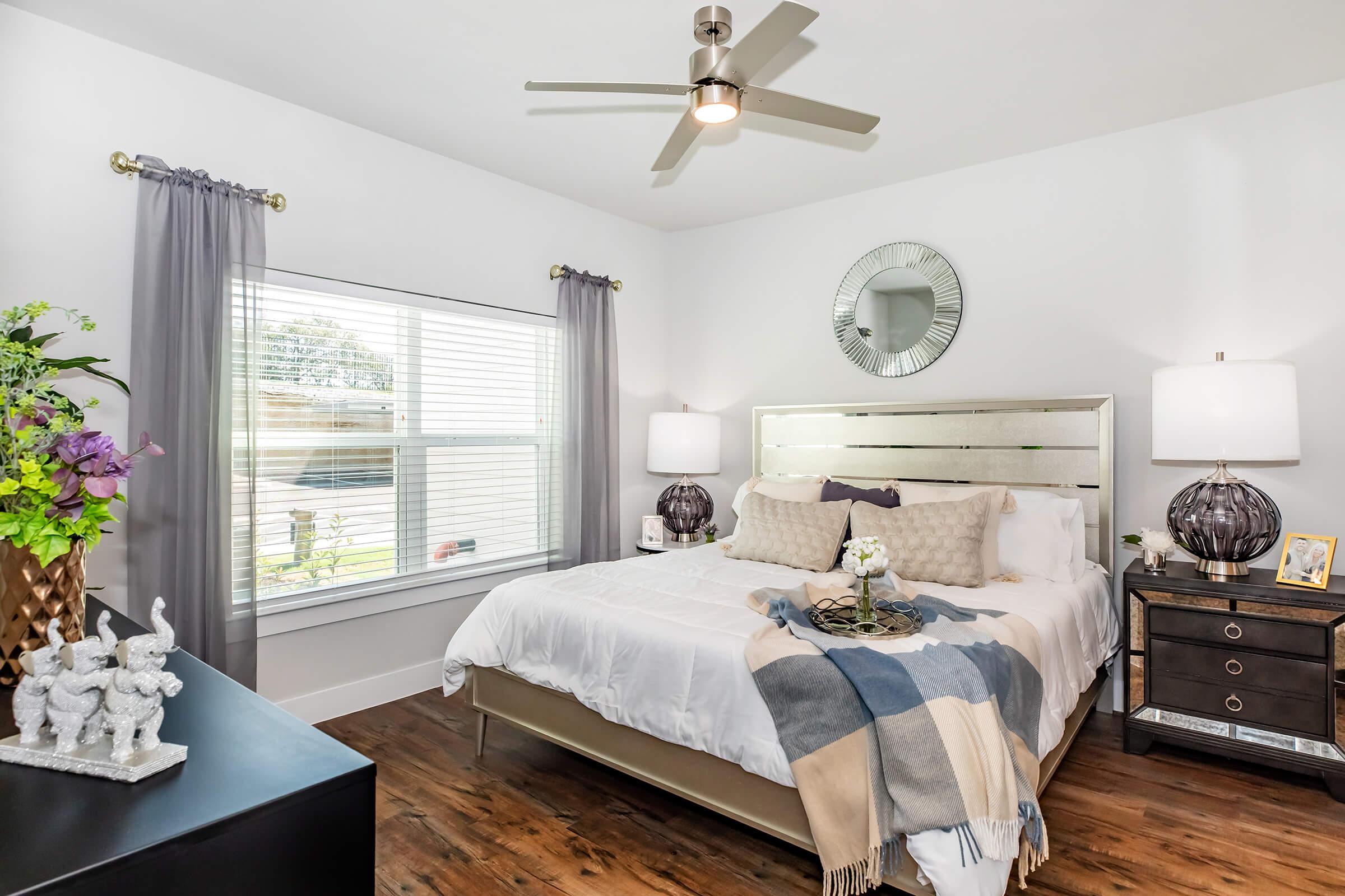 A tastefully decorated bedroom featuring a white bed with patterned pillows, a gray and blue throw blanket, two lamps on nightstands, and a large window with sheer curtains. A round mirror hangs above the headboard, and a decorative vase with flowers adds a touch of color. The hardwood floor complements the modern decor.